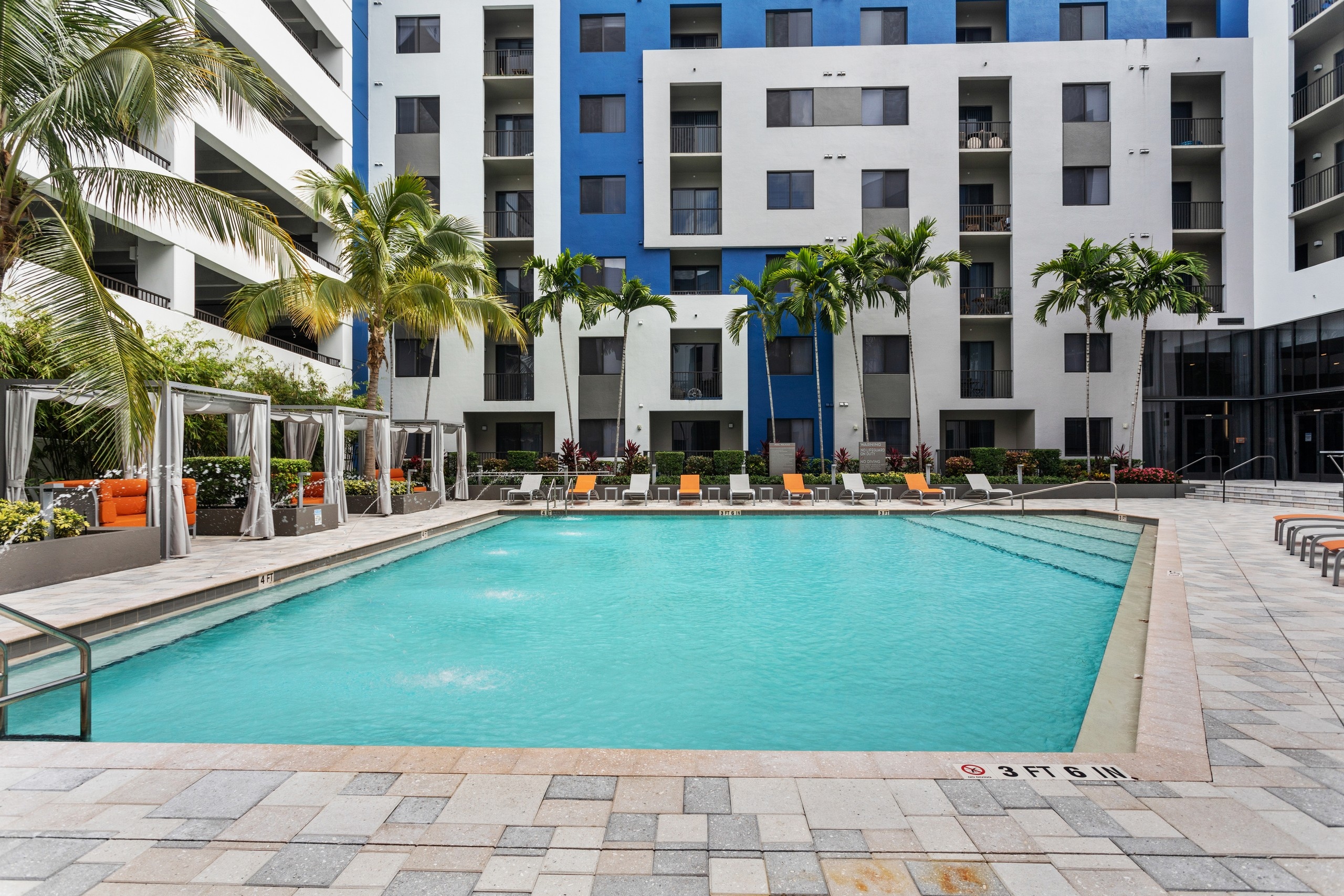 Daytime view of the AMLI 8800 pool with fountain water feature and orange and gray lounge chairs with cabanas and palm trees
