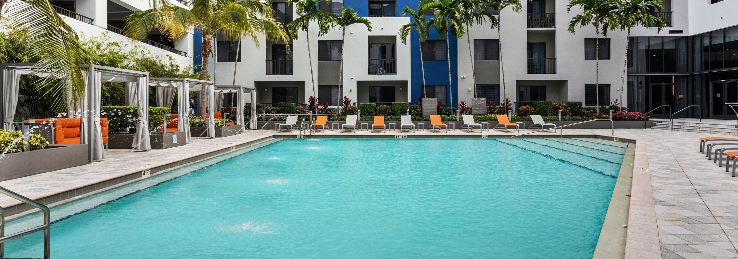 Daytime view of the AMLI 8800 pool with fountain water feature and orange and gray lounge chairs with cabanas and palm trees