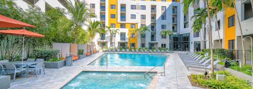 Daytime view of swimming pool and square heated spa at AMLI 8800 surrounded by lush landscape, umbrellas and pool furniture
