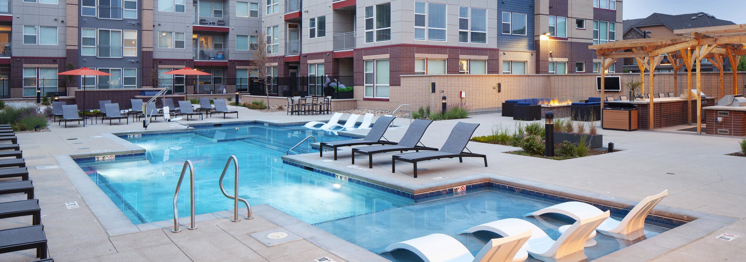 Evening view at AMLI Littleton Village pool with lounge seating and fire pit with TV and grill and apartment building in rear
