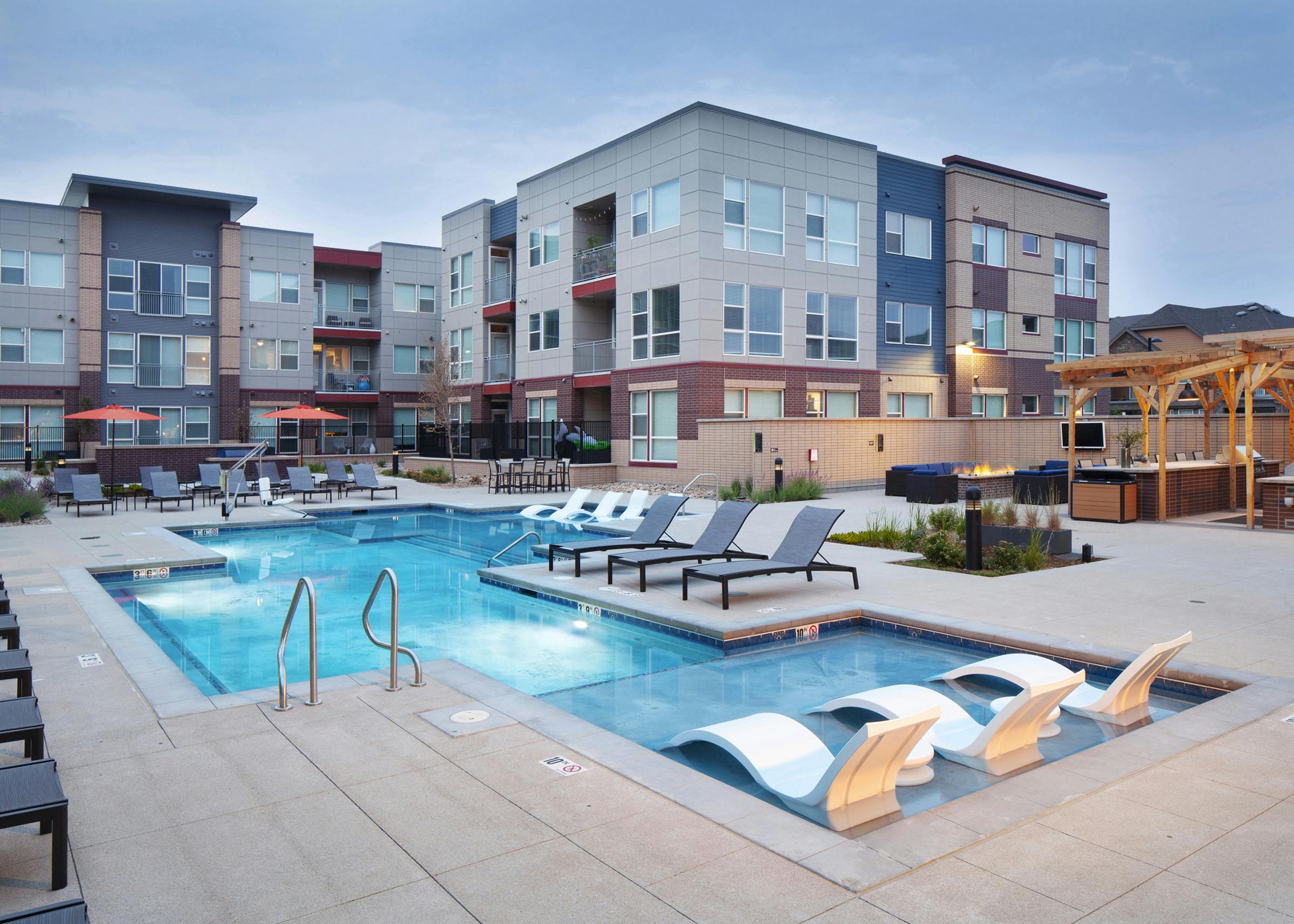 Evening view at AMLI Littleton Village pool with lounge seating and fire pit with TV and grill and apartment building in rear