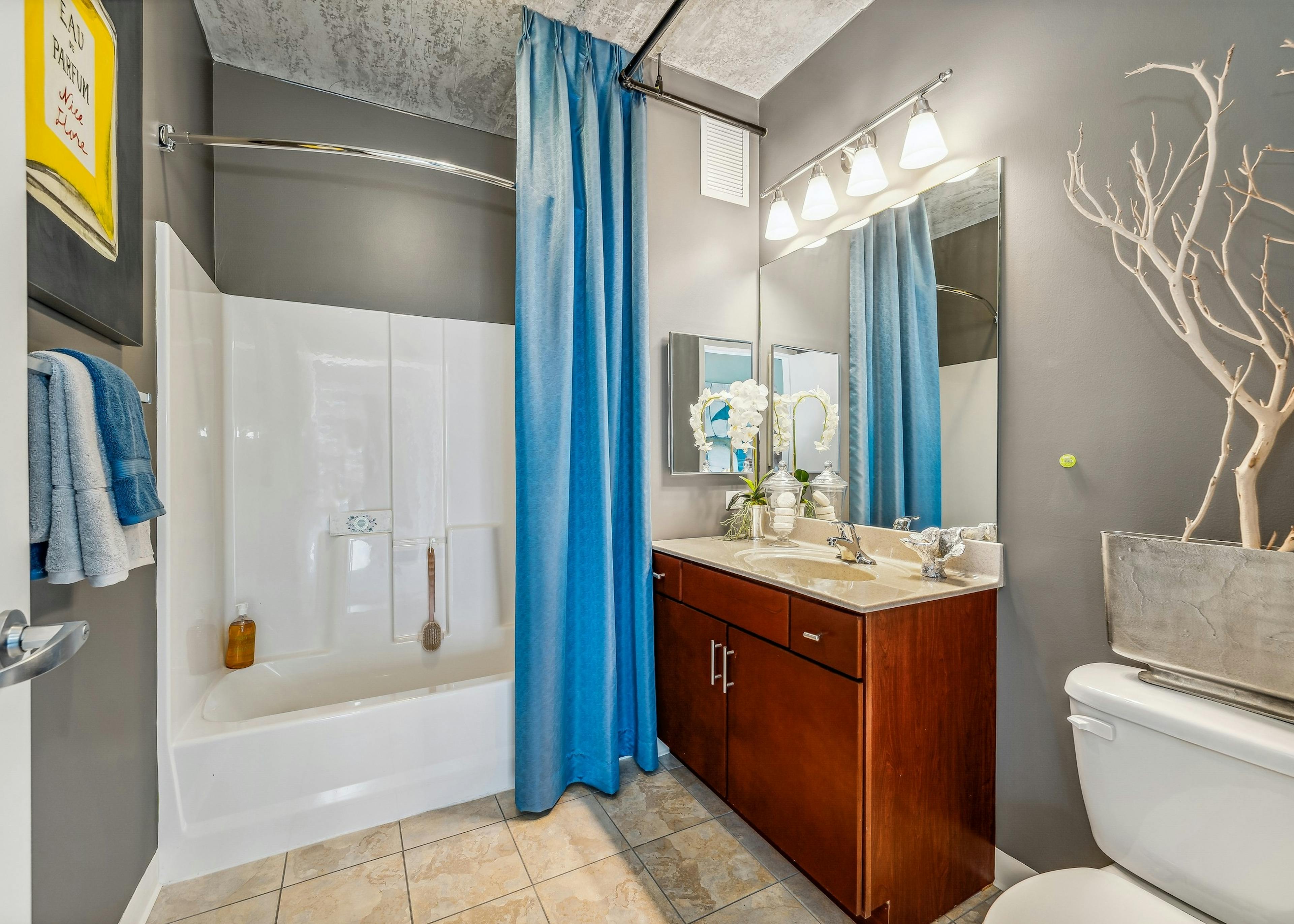 Bathroom at AMLI 900 with dark wood vanity with large mirror and white tub with blue shower curtain and gray walls