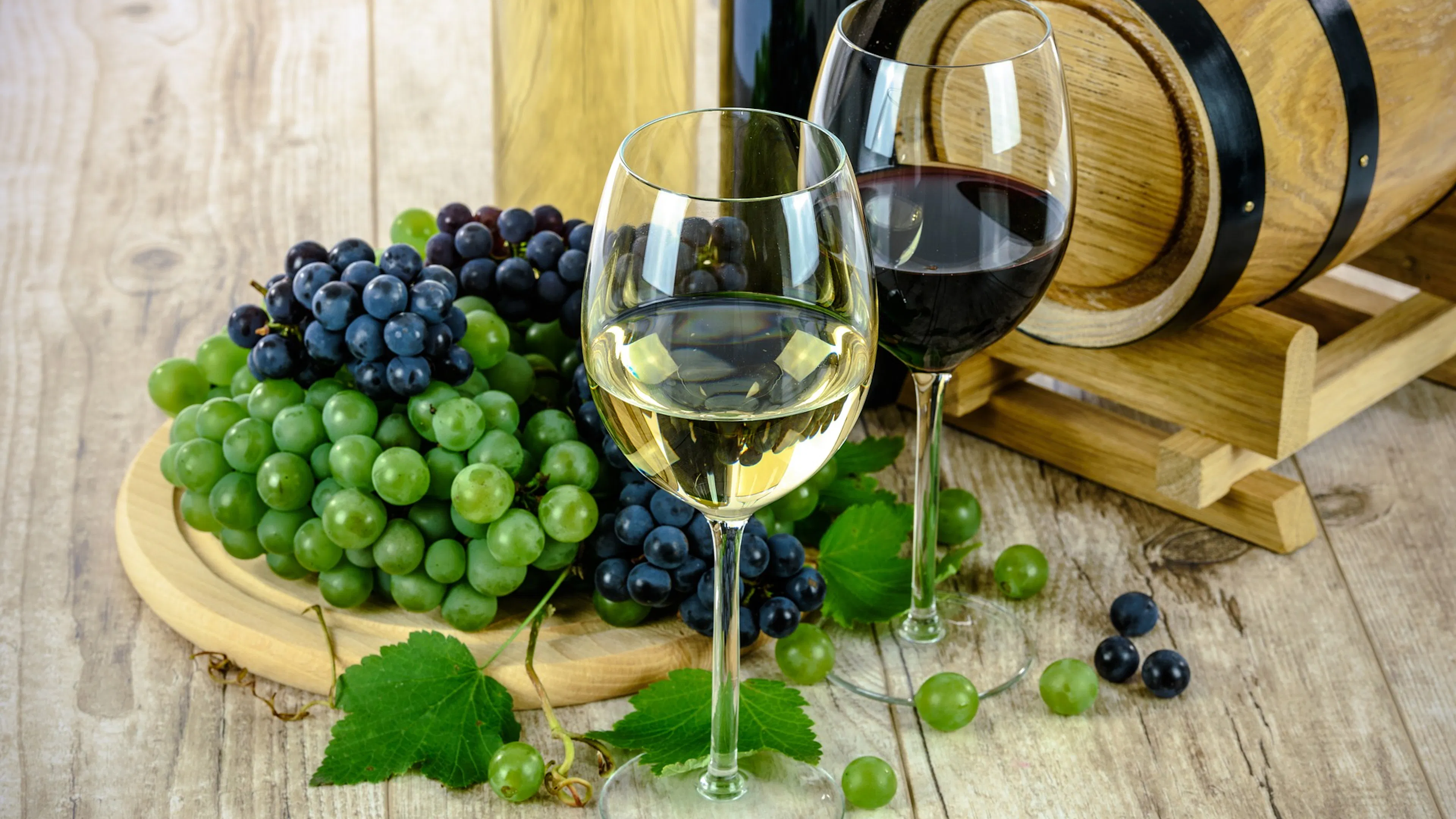 Two wine glasses with white and red wine next to bunches of red and green grapes on a wooden serving board and table