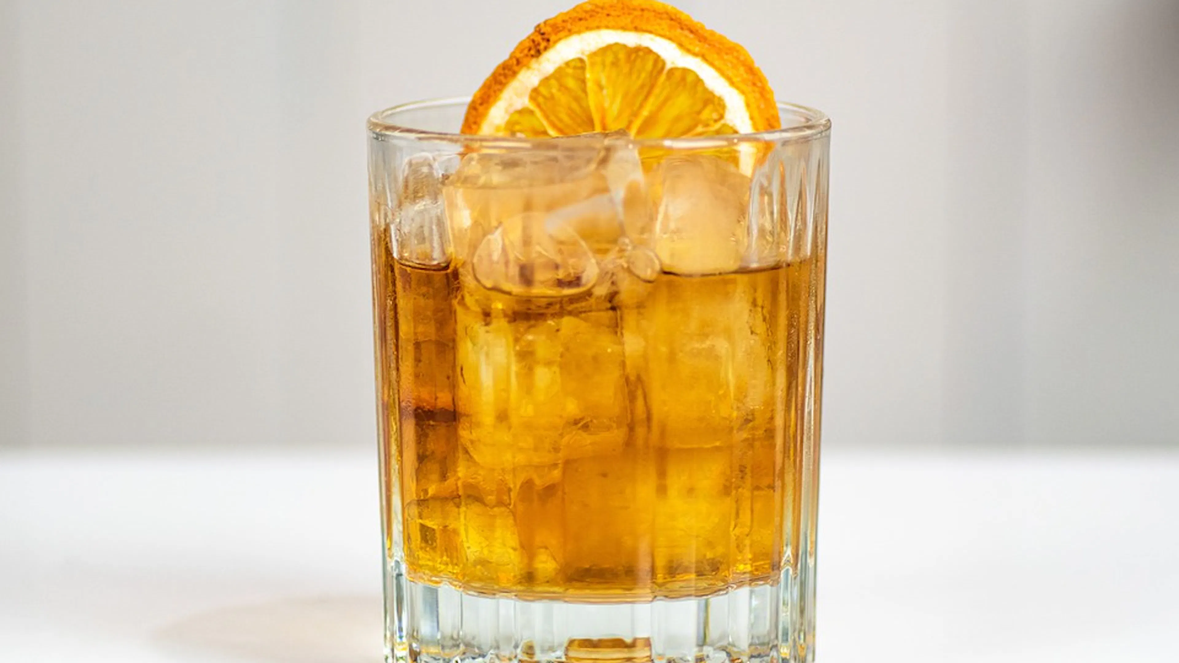 An old fashioned cocktail with an ice cube and orange wedge against a light background