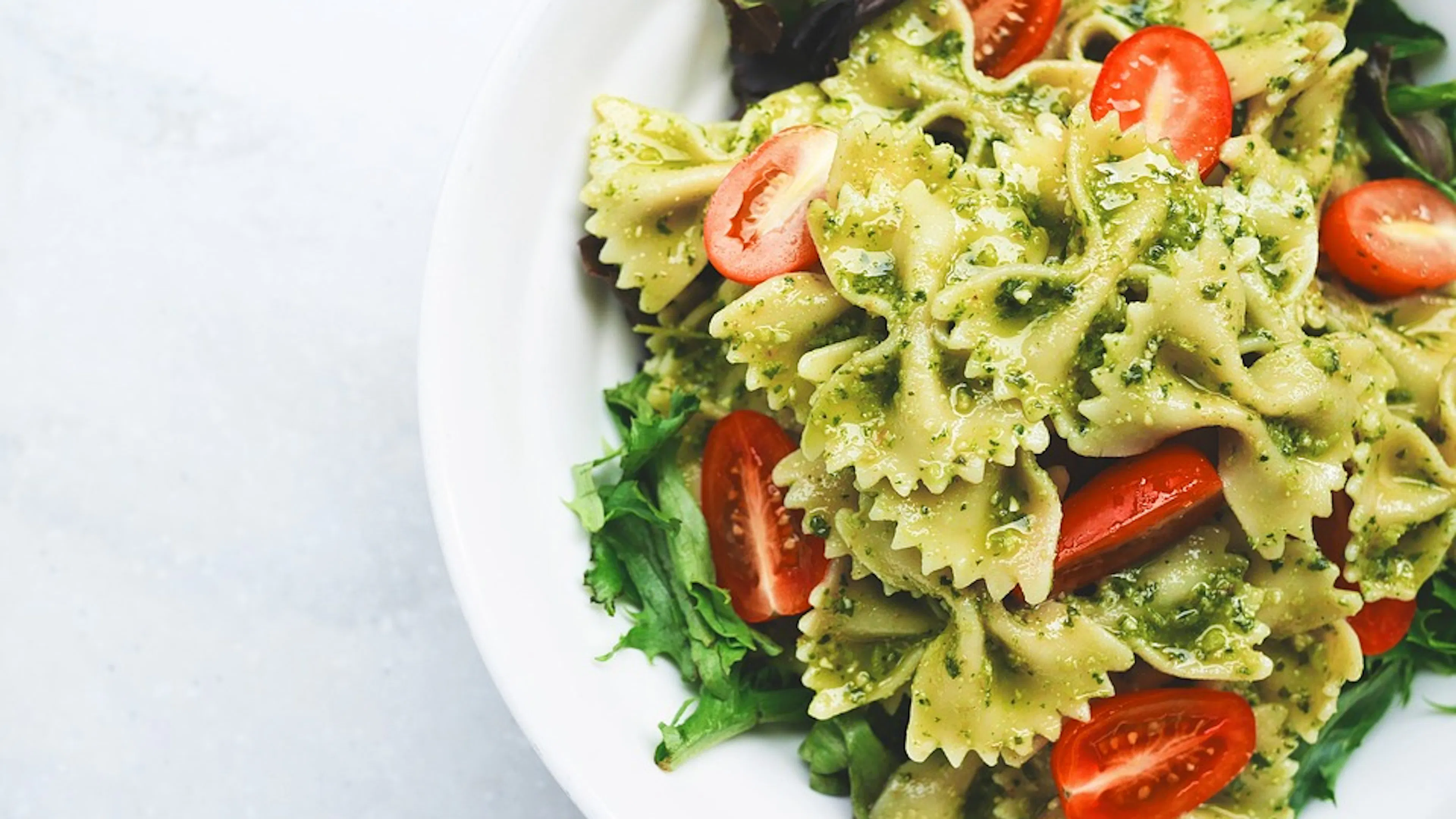 A white bowl filled with pesto farfalle pasta and chopped cherry tomatoes on a bed of greens