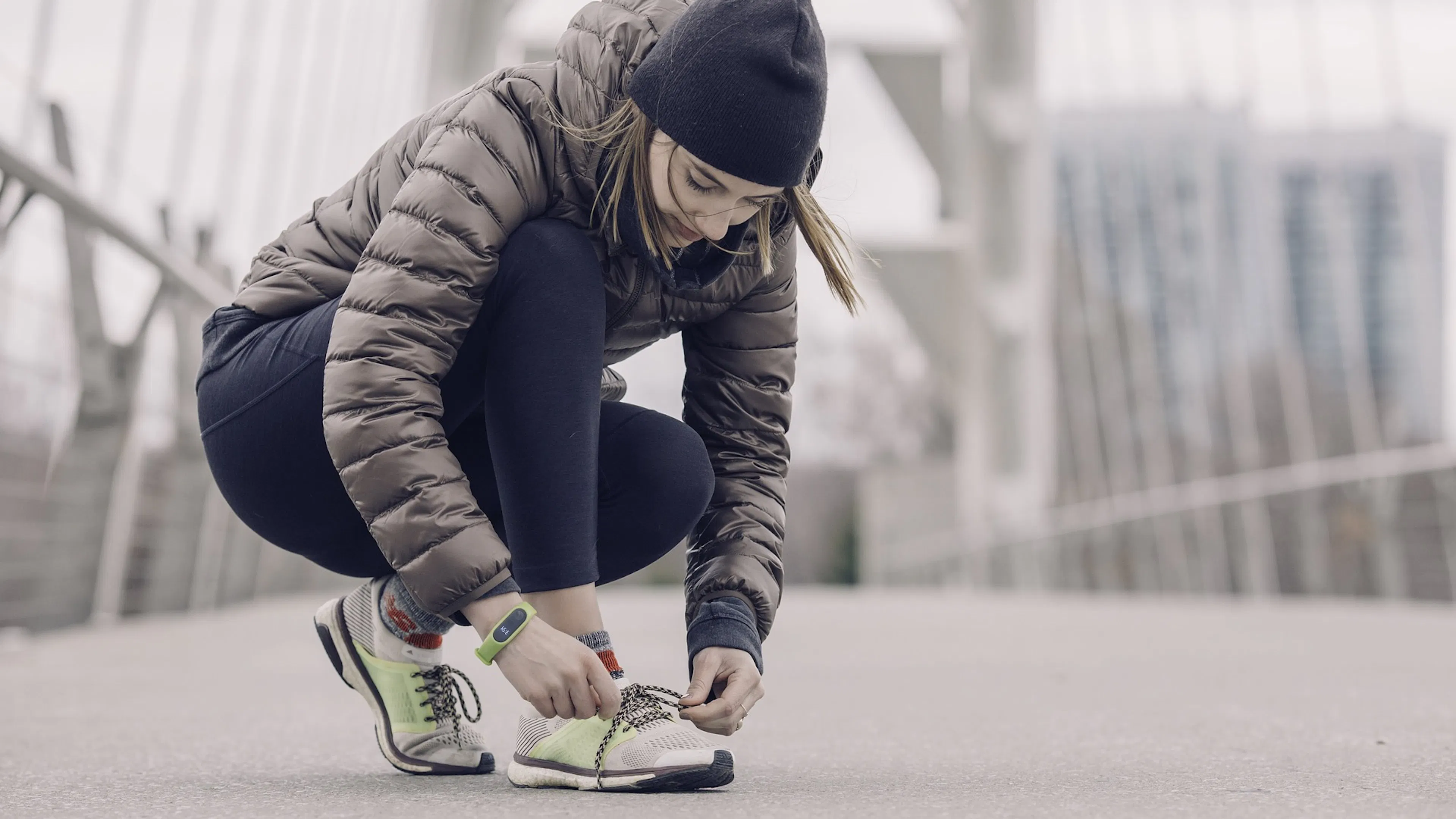 young woman in black hat with puffy jacket tying white and green sneakers while she kneels on a bridge