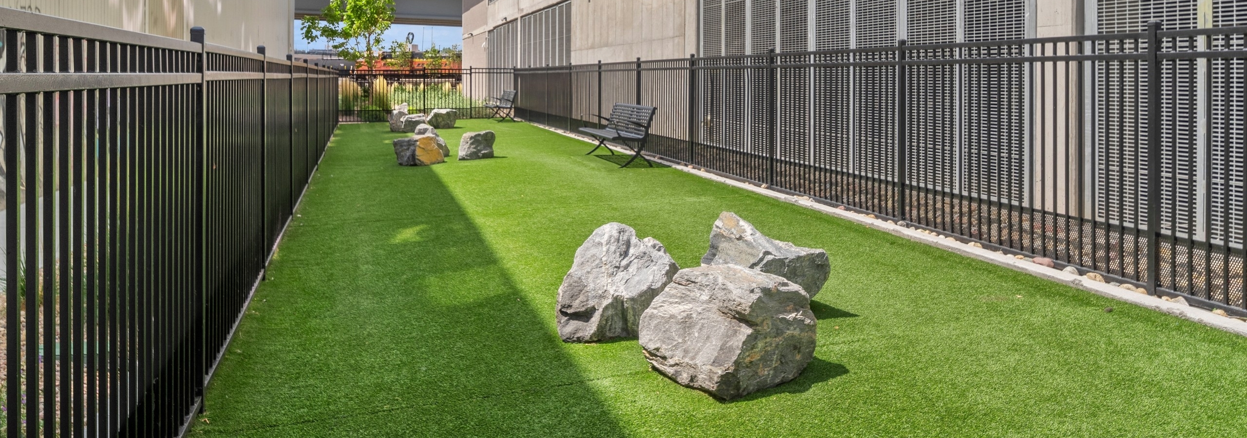 Fenced in Paw park at AMLI RiNo apartments between 2 tan building with grass and large boulders and 2 benches