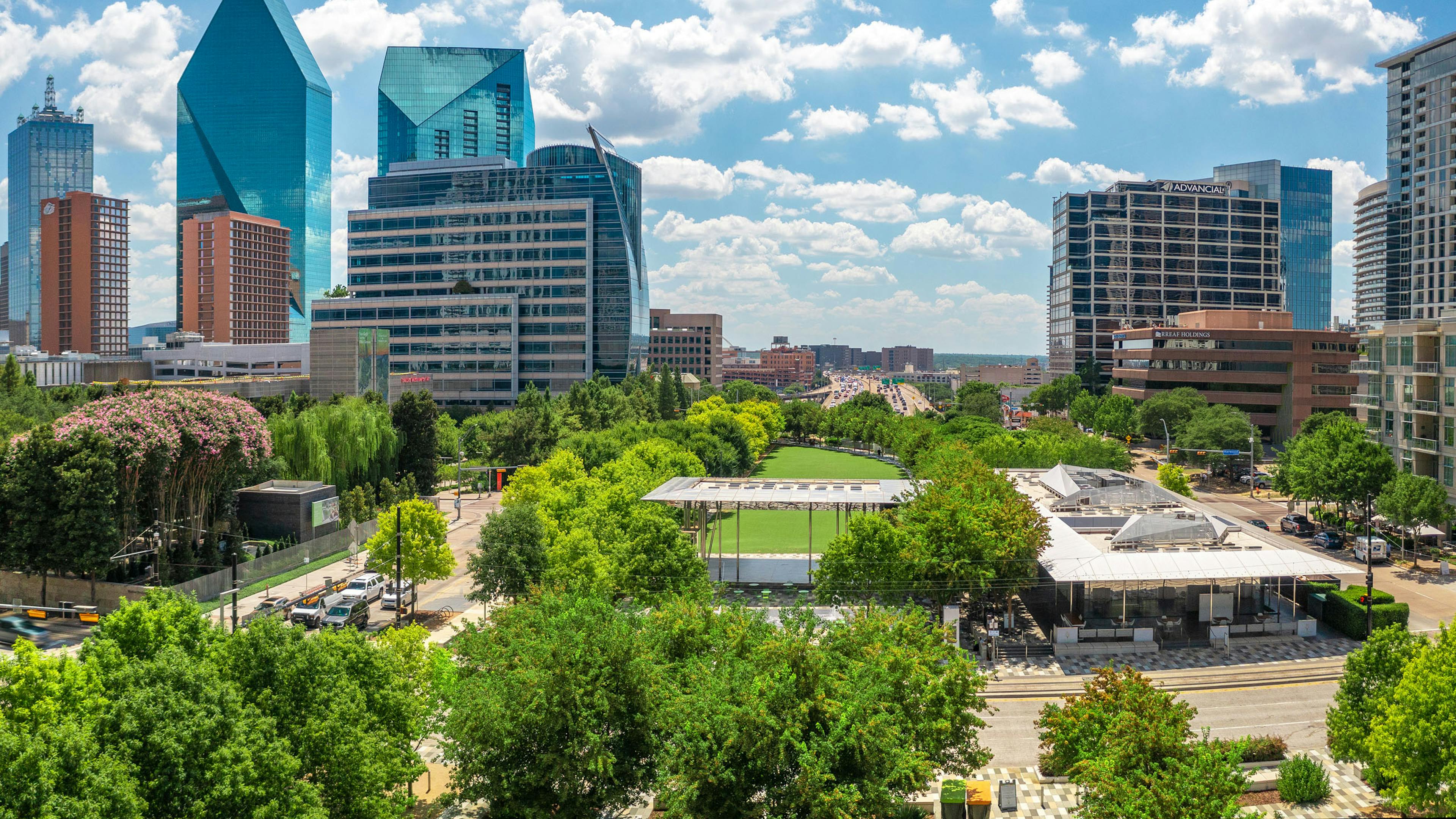 Aerial view of Klyde Warren Park with lush green trees circling grassy area and high rise buildings all around and blue sky