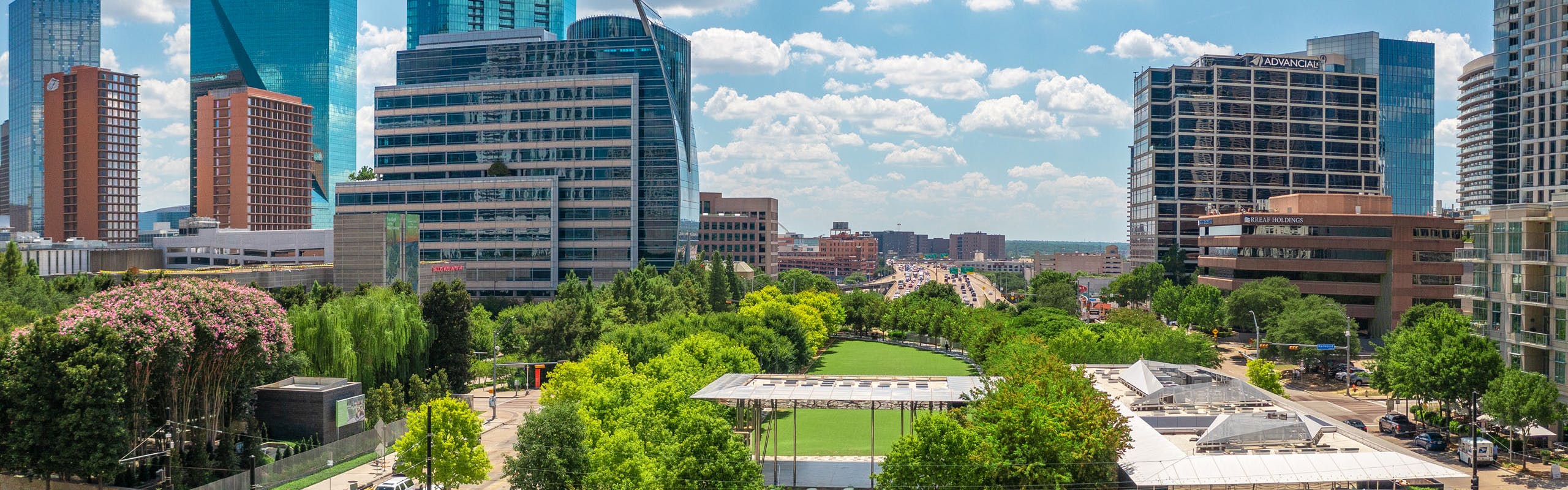 Aerial view of Klyde Warren Park with lush green trees circling grassy area and high rise buildings all around and blue sky