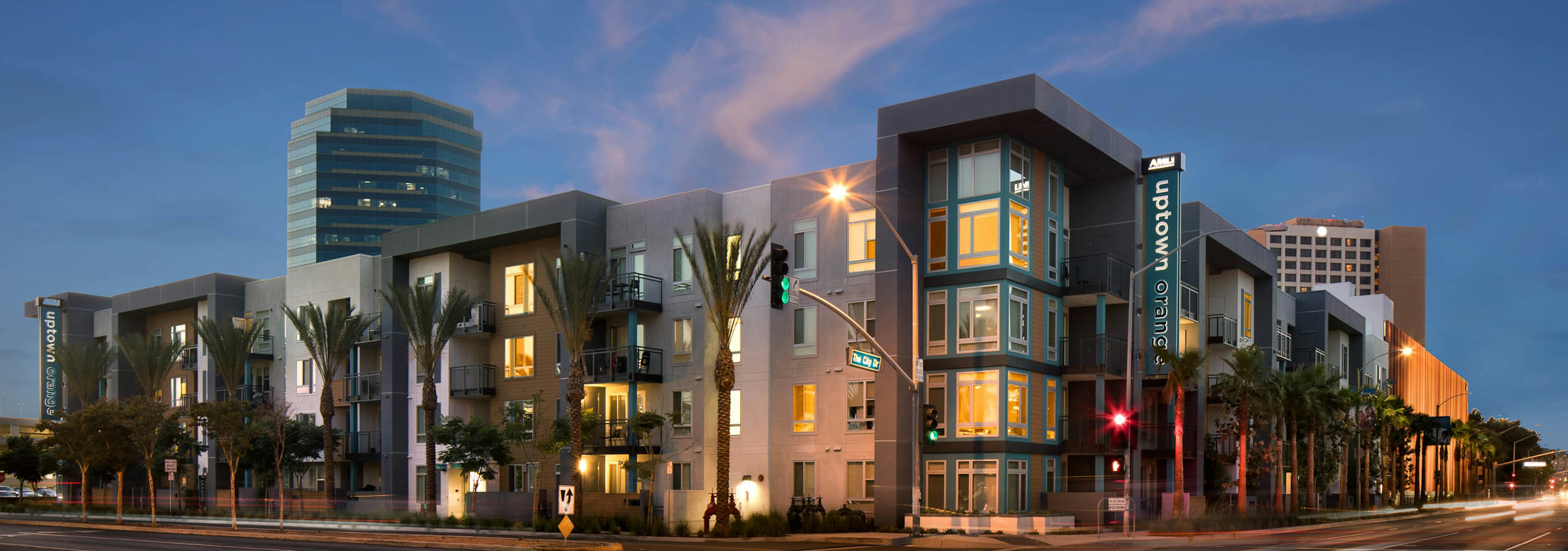 Exterior rendering of four story AMLI Uptown Orange apartment building at dusk surrounded by palm trees and lit monument