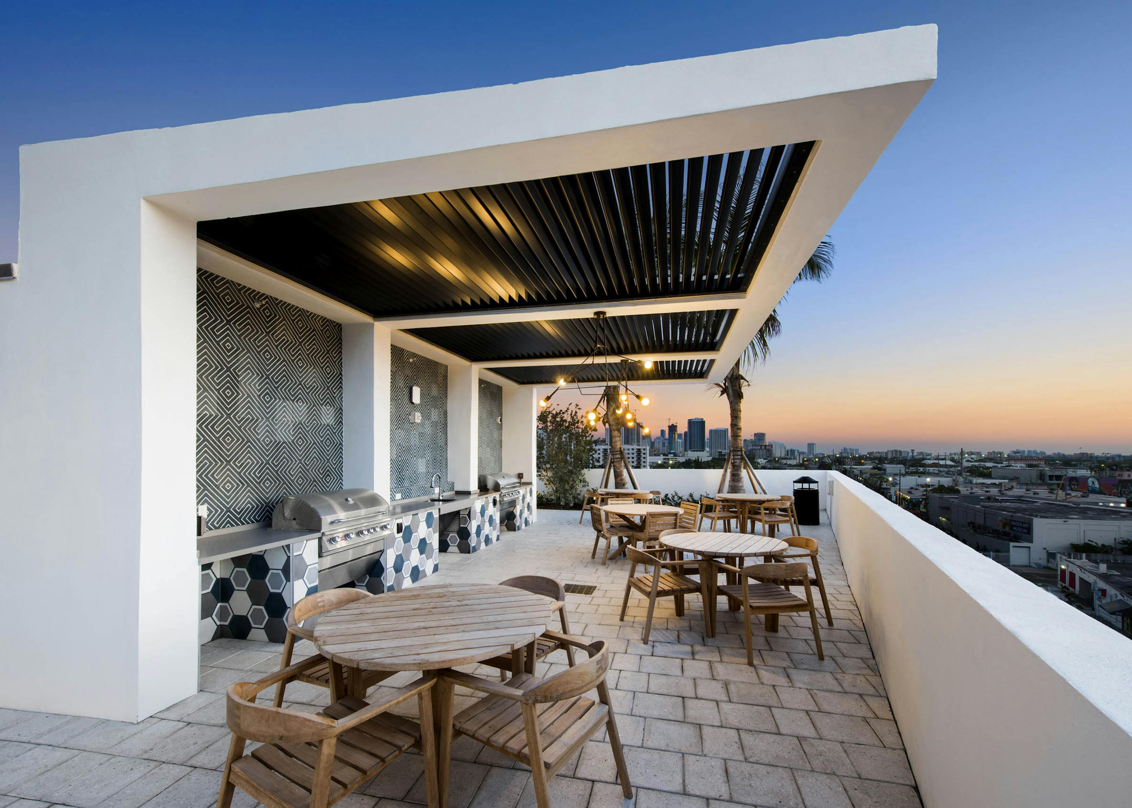 Dusk view of rooftop grill stations with slatted overhang and dining tables along short white wall and view of the city