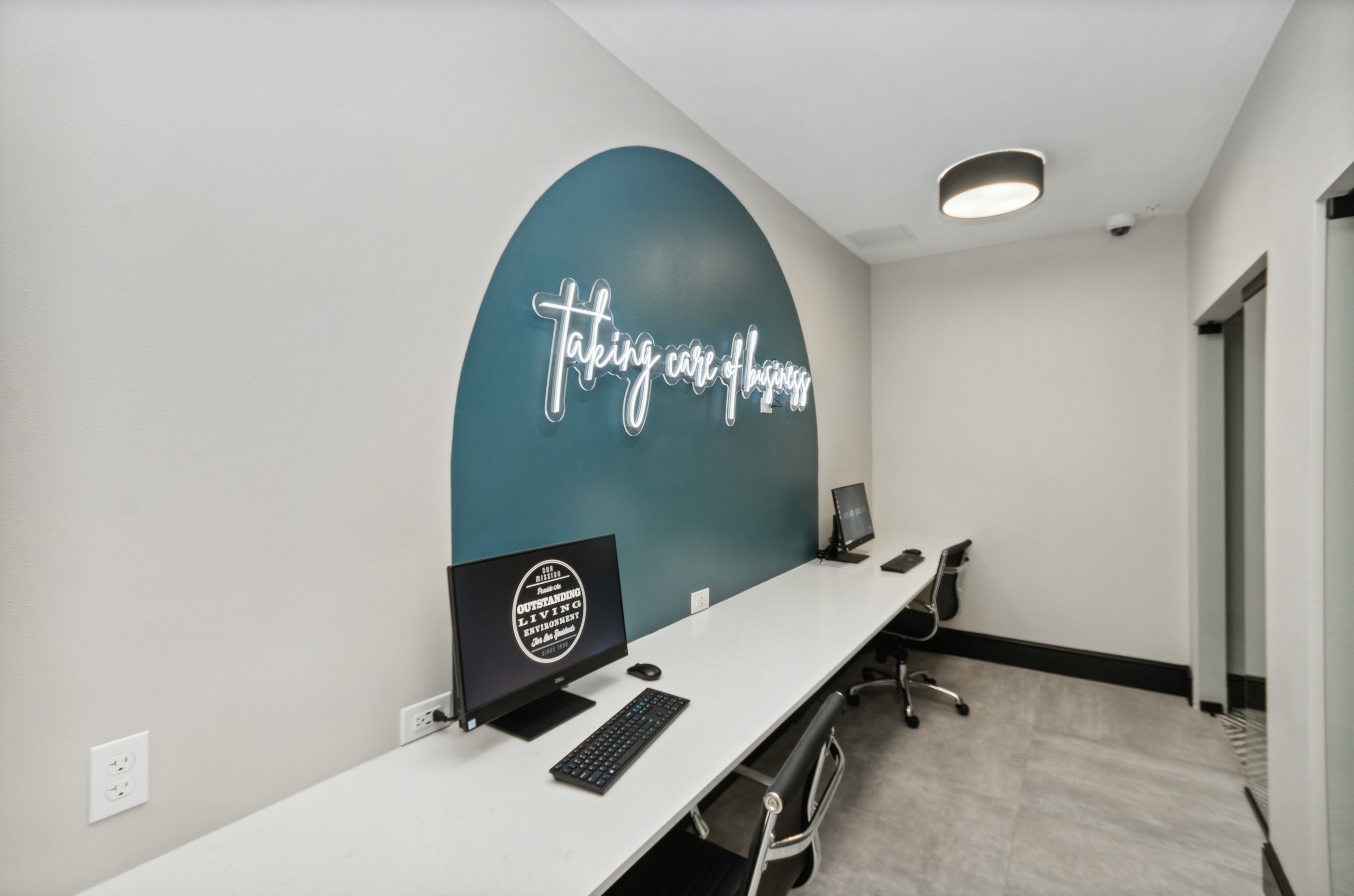 Business center at AMLI Campion Trail with 2 computers on long white desk and two black chairs and wall with white neon sign