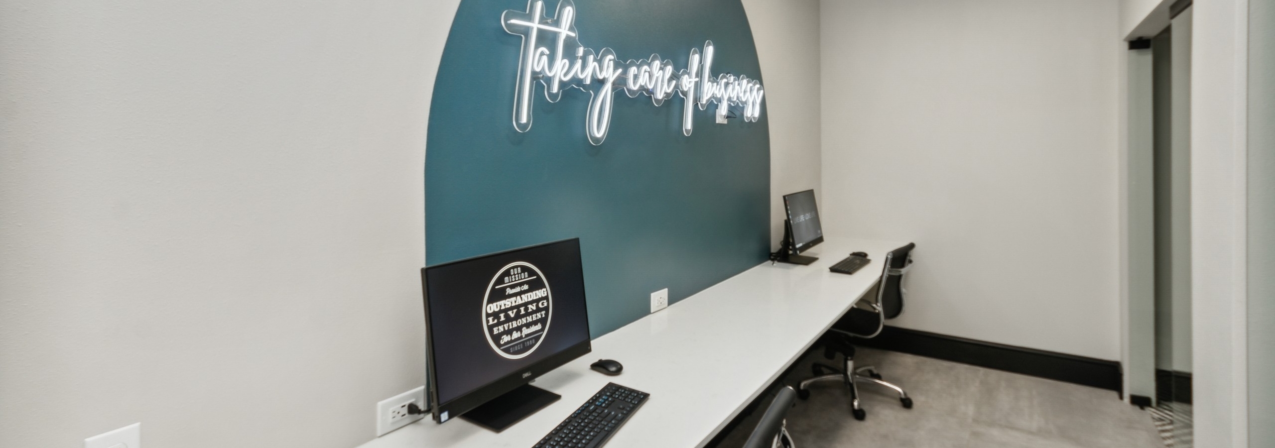 Business center at AMLI Campion Trail with 2 computers on long white desk and two black chairs and wall with white neon sign