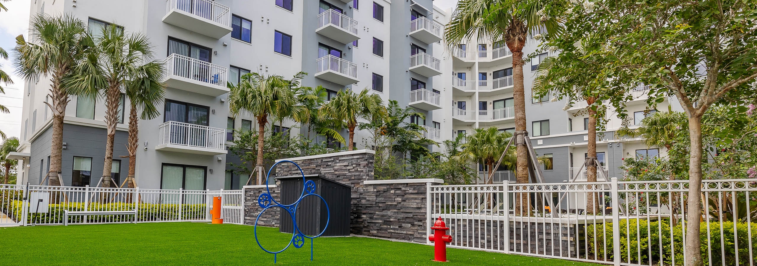 Exterior view of AMLI Park West apartments dog park with faux green grass and agility equipment and an orange water station