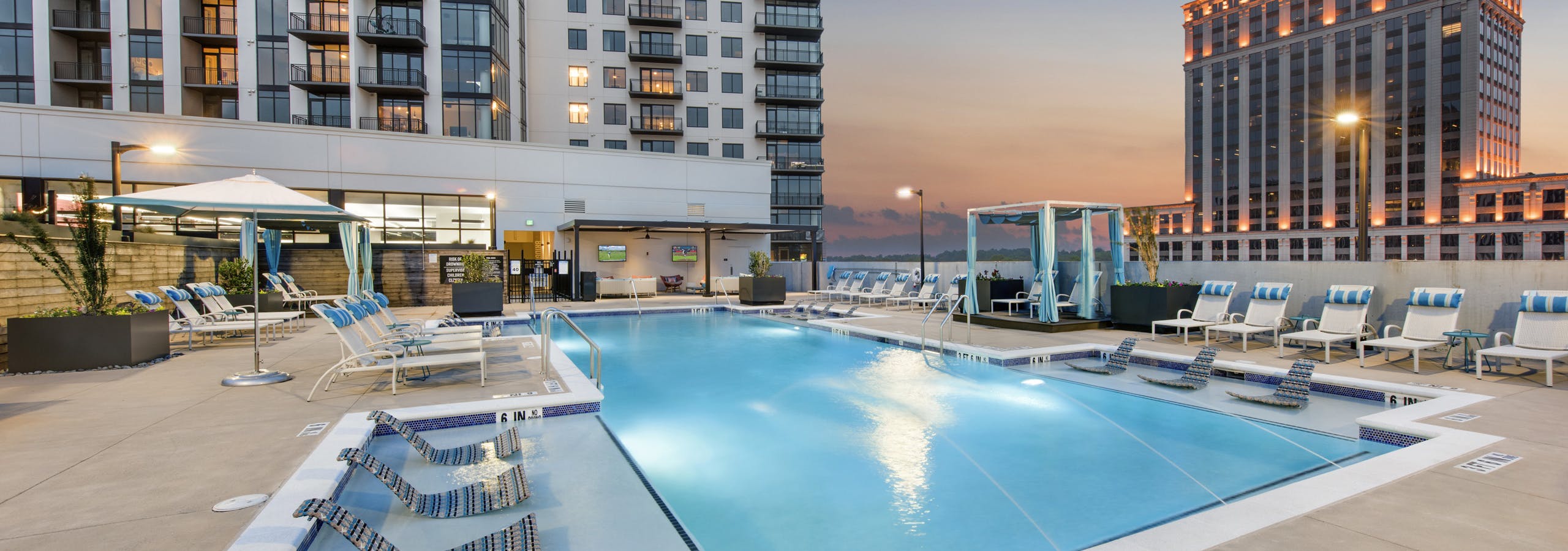 Dusk view of pool area at AMLI Lenox apartment building with lounge chairs and cabanas around the pool with building set back