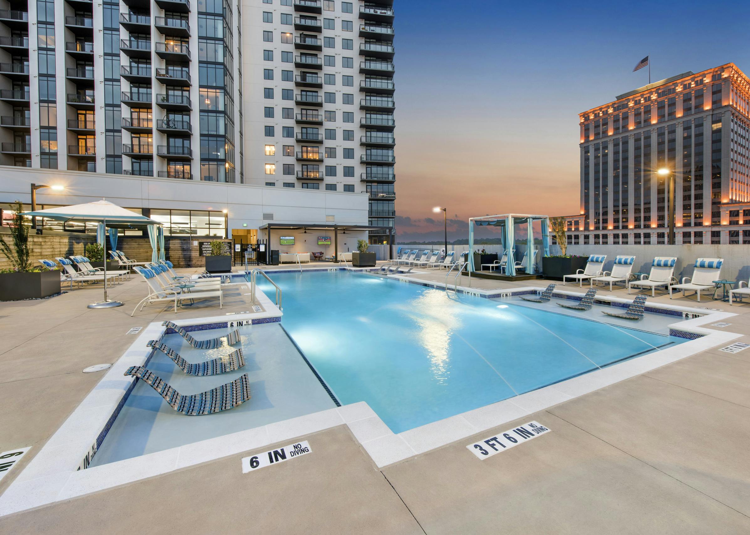 Dusk view of pool area at AMLI Lenox apartment building with lounge chairs and cabanas around the pool with building set back