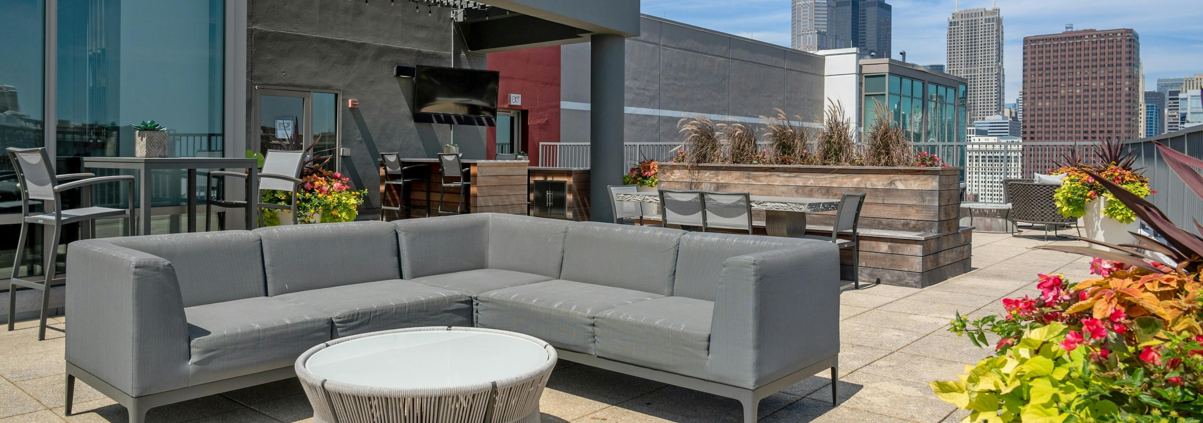 AMLI 900 rooftop with grey sectional couch and round table on concrete patio with wood planters and flowers and city skyline view