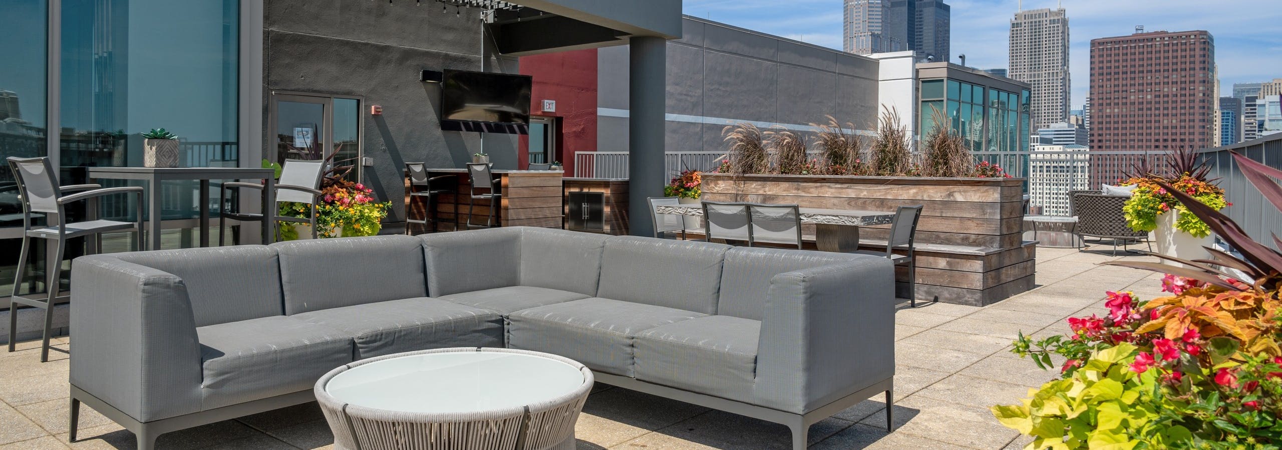 AMLI 900 rooftop with grey sectional couch and round table on concrete patio with wood planters and flowers and city skyline view