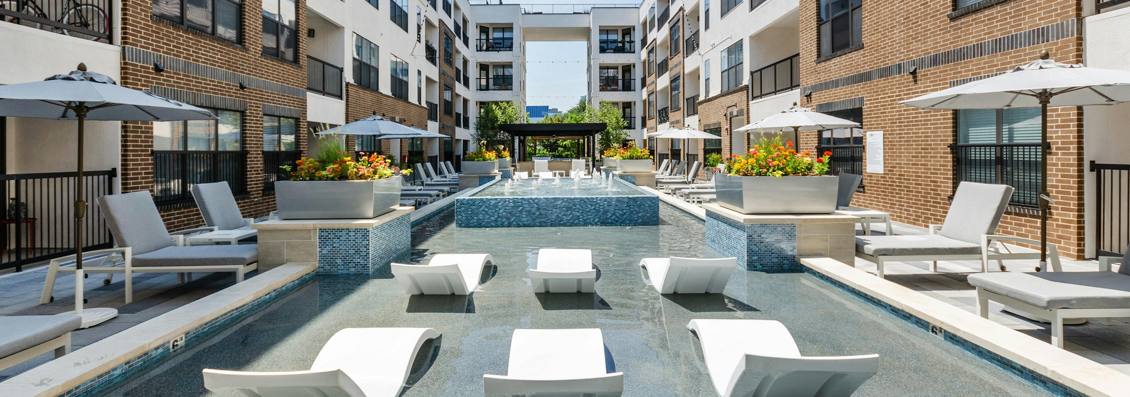 A sunlit shallow pool at AMLI Quadrangle apartments with lounge seating with shading umbrellas and flowers in metal planters