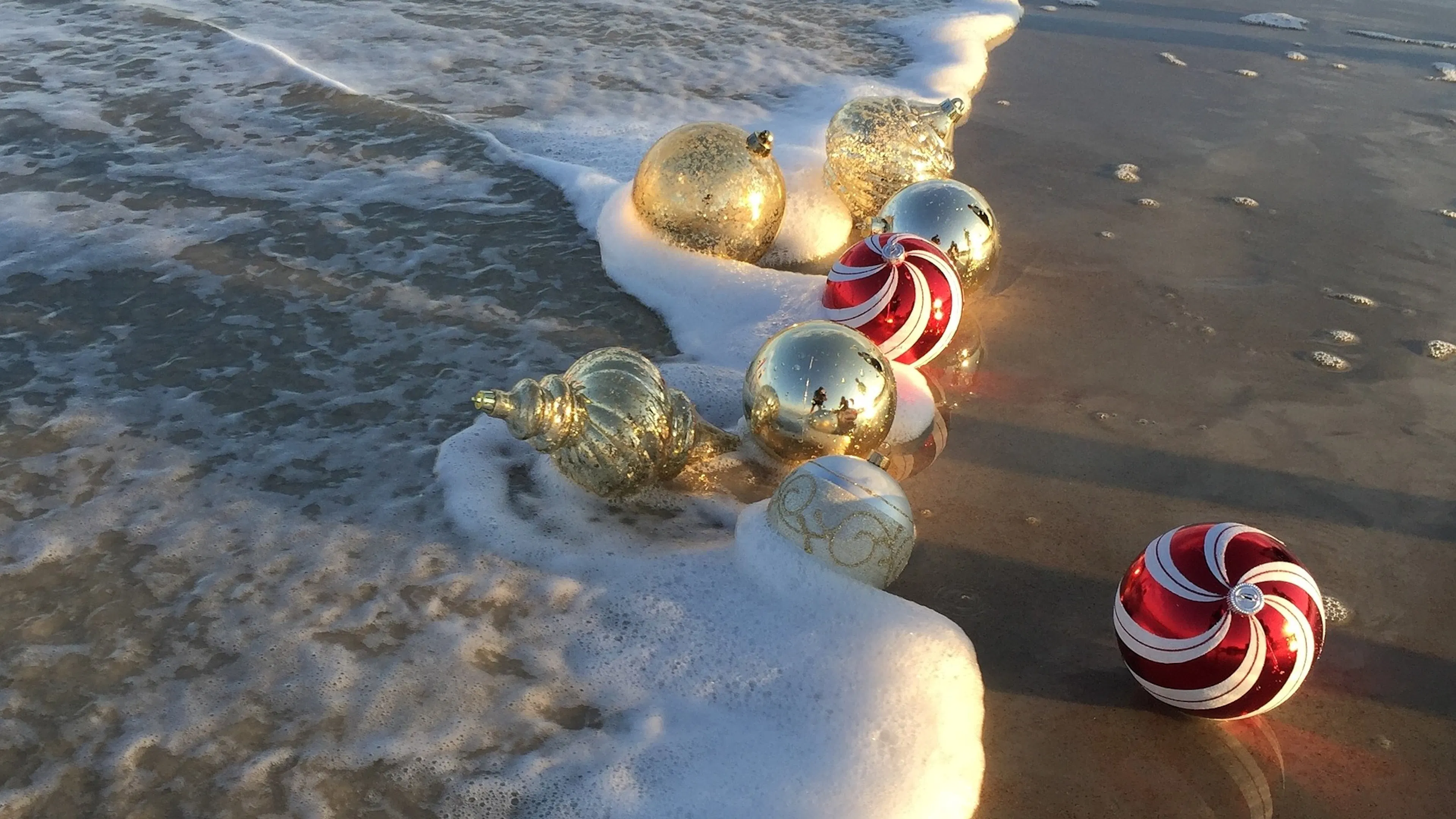 Glittery gold and candy stripe red Christmas ornaments on the beach with the tide washing up to them