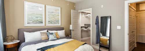 Interior of AMLI West Plano bedroom with tan walls and a white bed with a yellow throw blanket and a peek into walk in closet