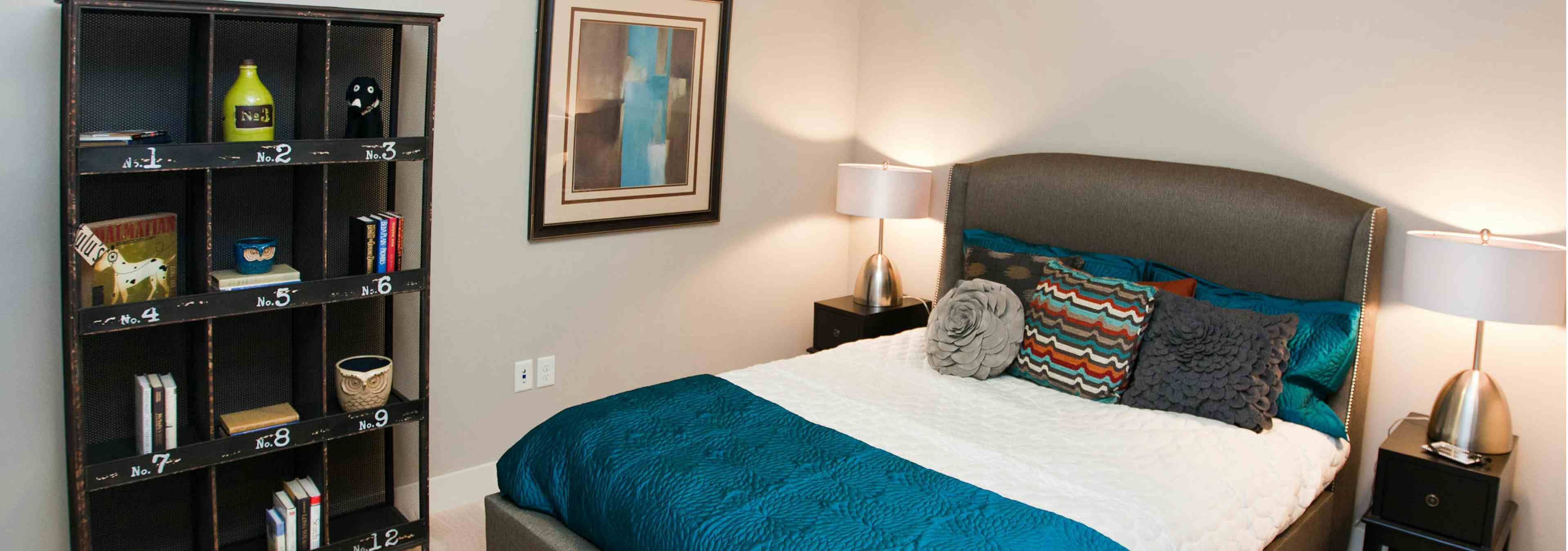 A bedroom at AMLI Riverfront Park apartments with a large bookcase side tables with lamps and artwork hanging on the wall