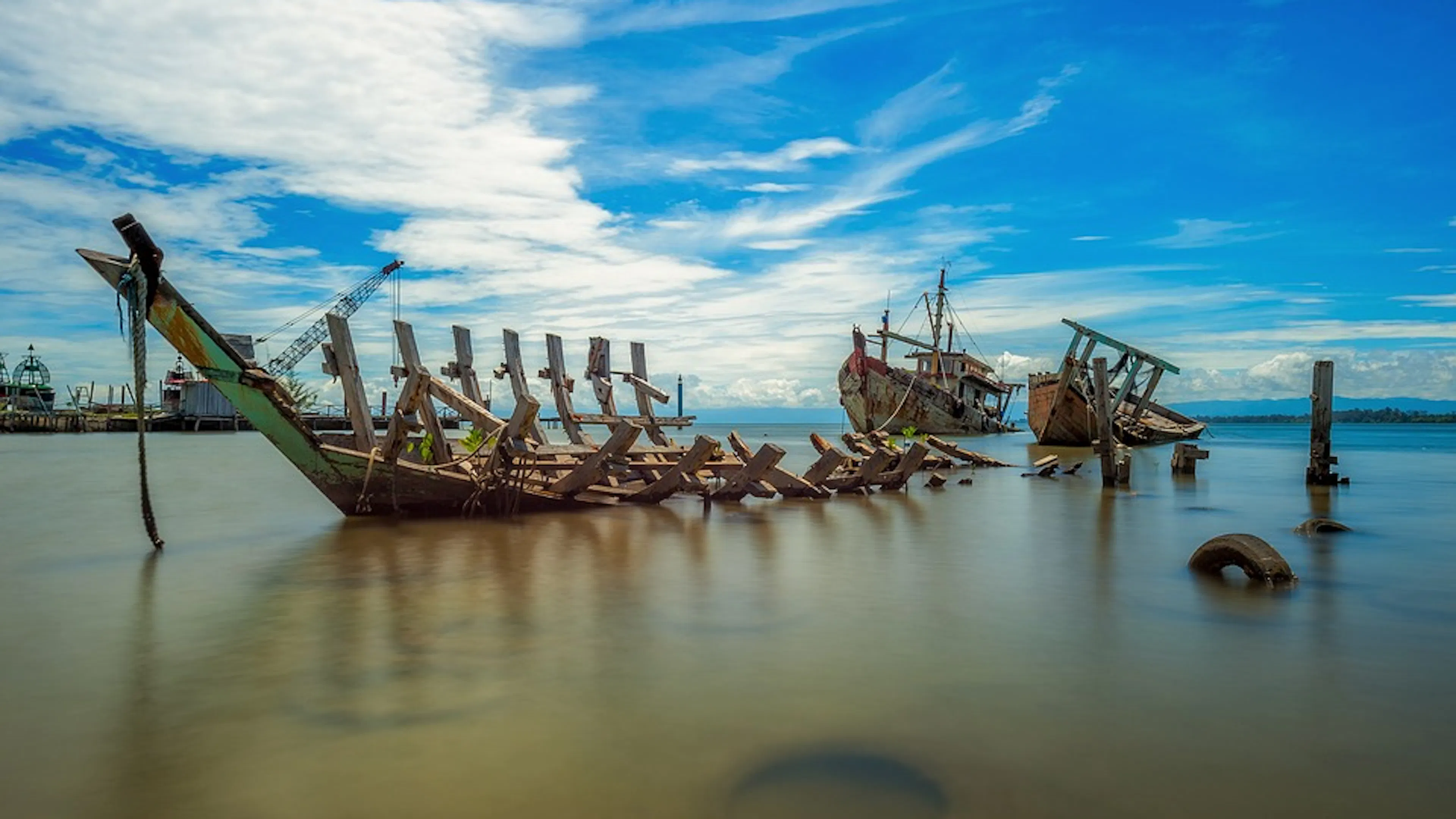 The metal hull of of a shipwrecked ship sitting at an angle in the shallow water of a shipyard