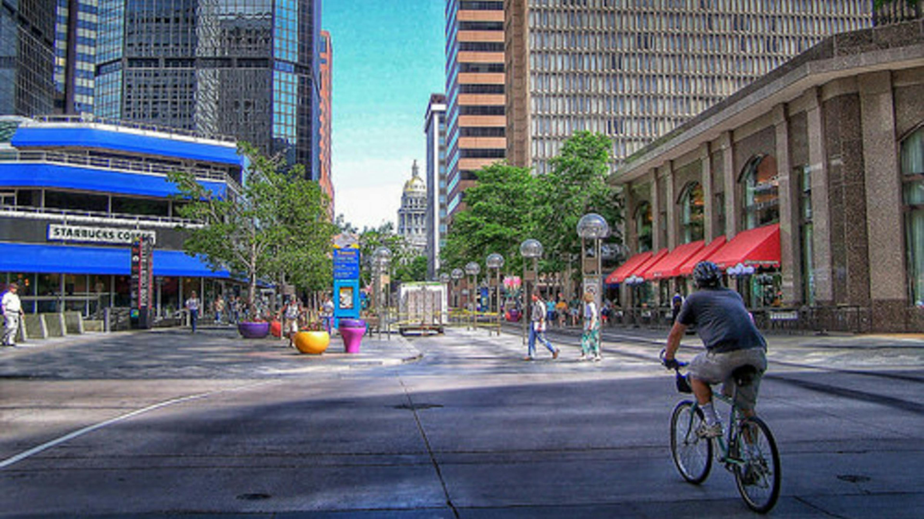 A view of the streets in downtown Denver with a person on a bike and people on the sidewalk and the Denver Capital Building