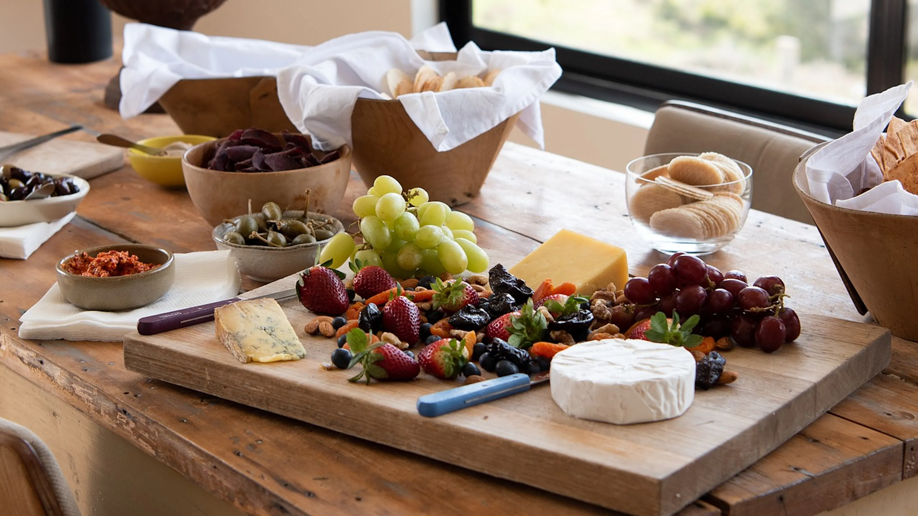 Wooden cutting board on a dining room table with an assortment of cheeses and crackers and fruits and condiments