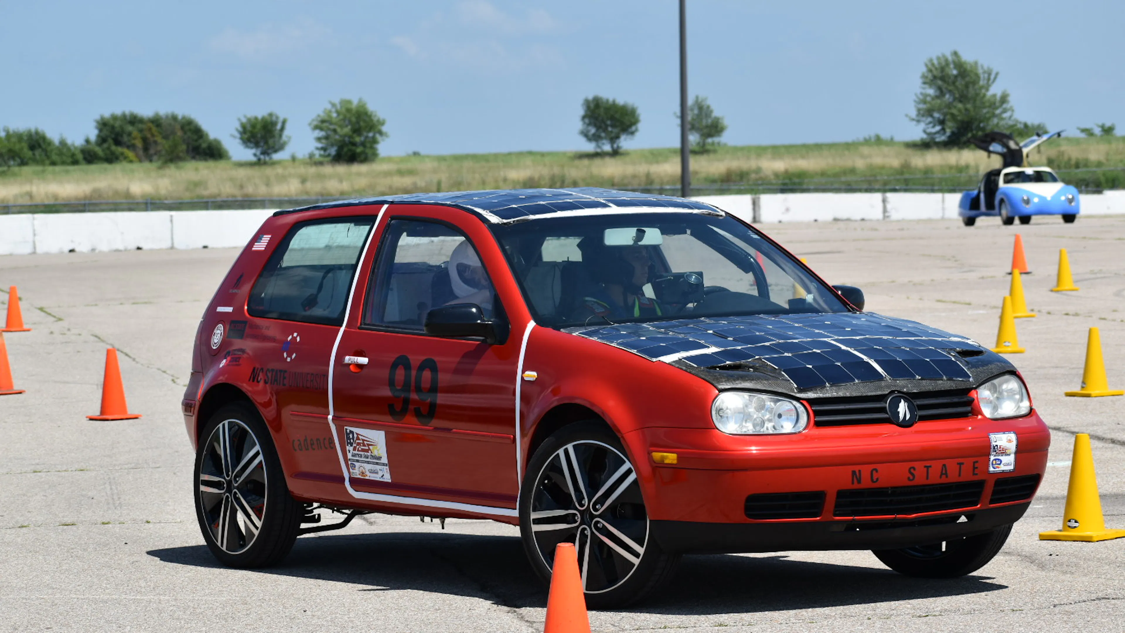 A red VW GTI hatchback with solar panels covering its roof and hood driving through a series of cones on a race track