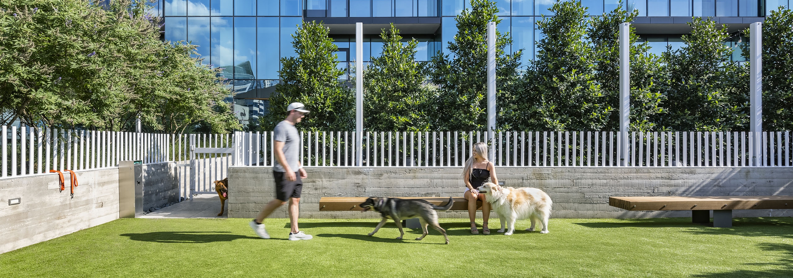 AMLI Fountain Place pet park with white fence and 2 people with dogs enjoying the park with building behind them