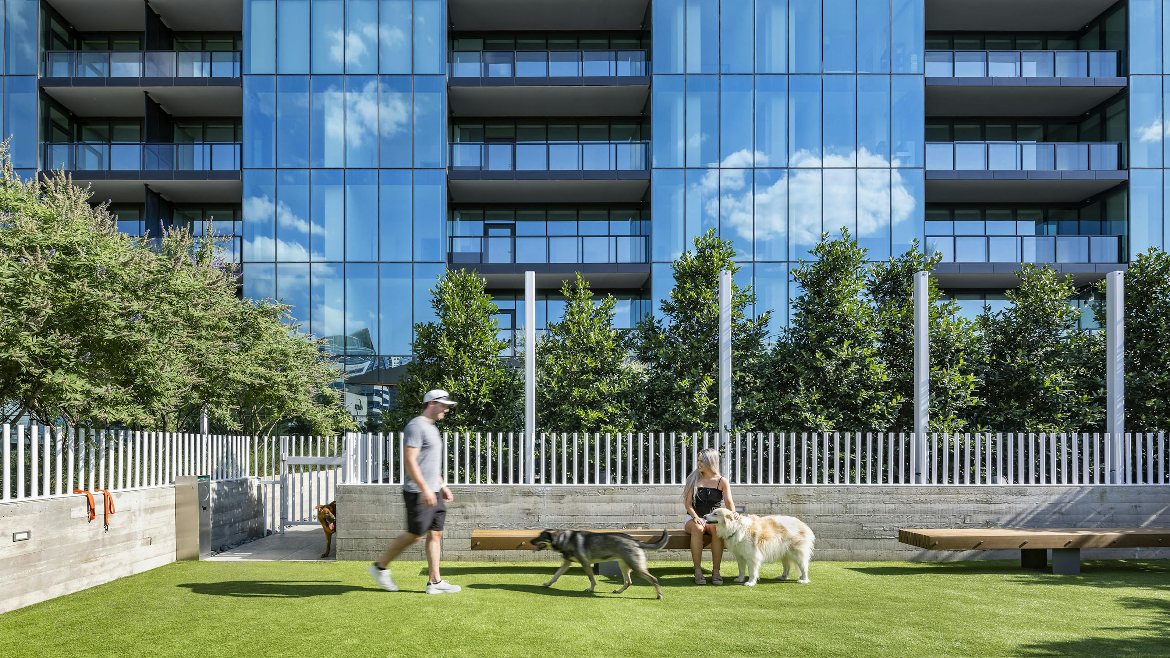 AMLI Fountain Place pet park with white fence and 2 people with dogs enjoying the park with building behind them
