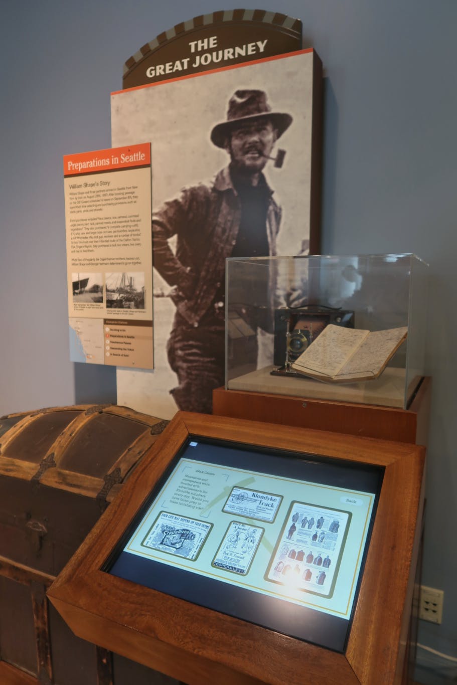 A digital informational plaque display alongside a physical exhibit on a gold miner's journey to the Yukon