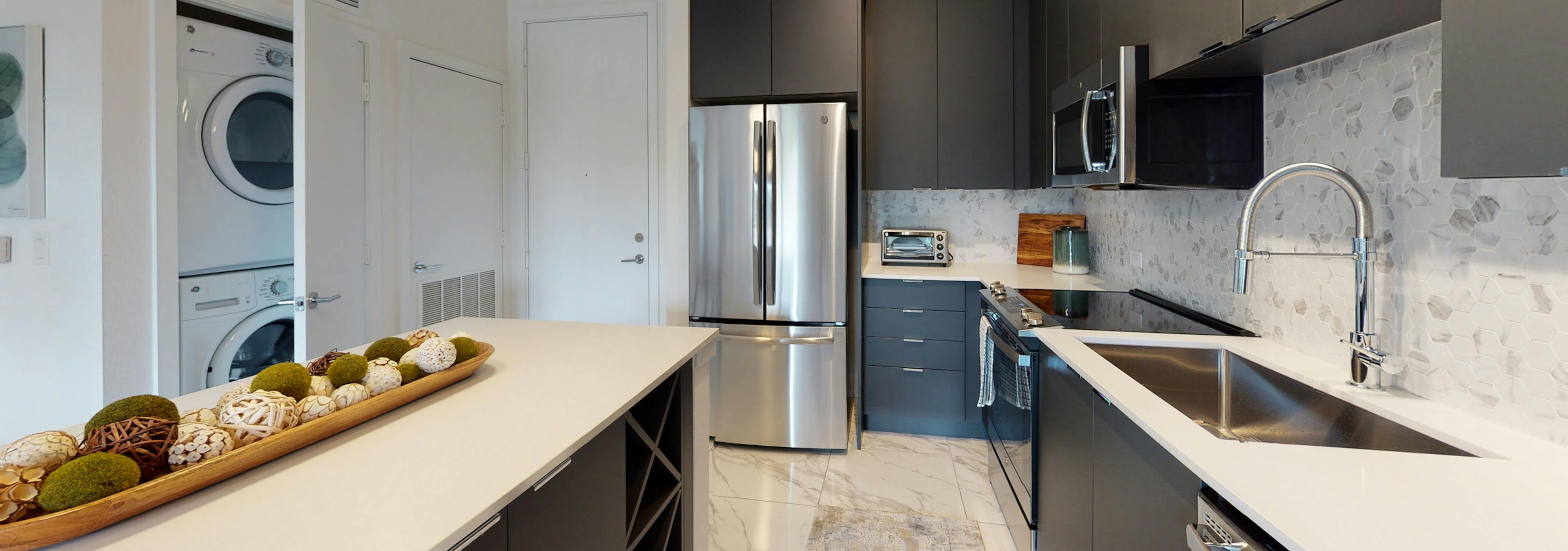 Kitchen at AMLI Park West apartments with dark cabinetry and white quartz counters and marble floors and stacked washer/dryer