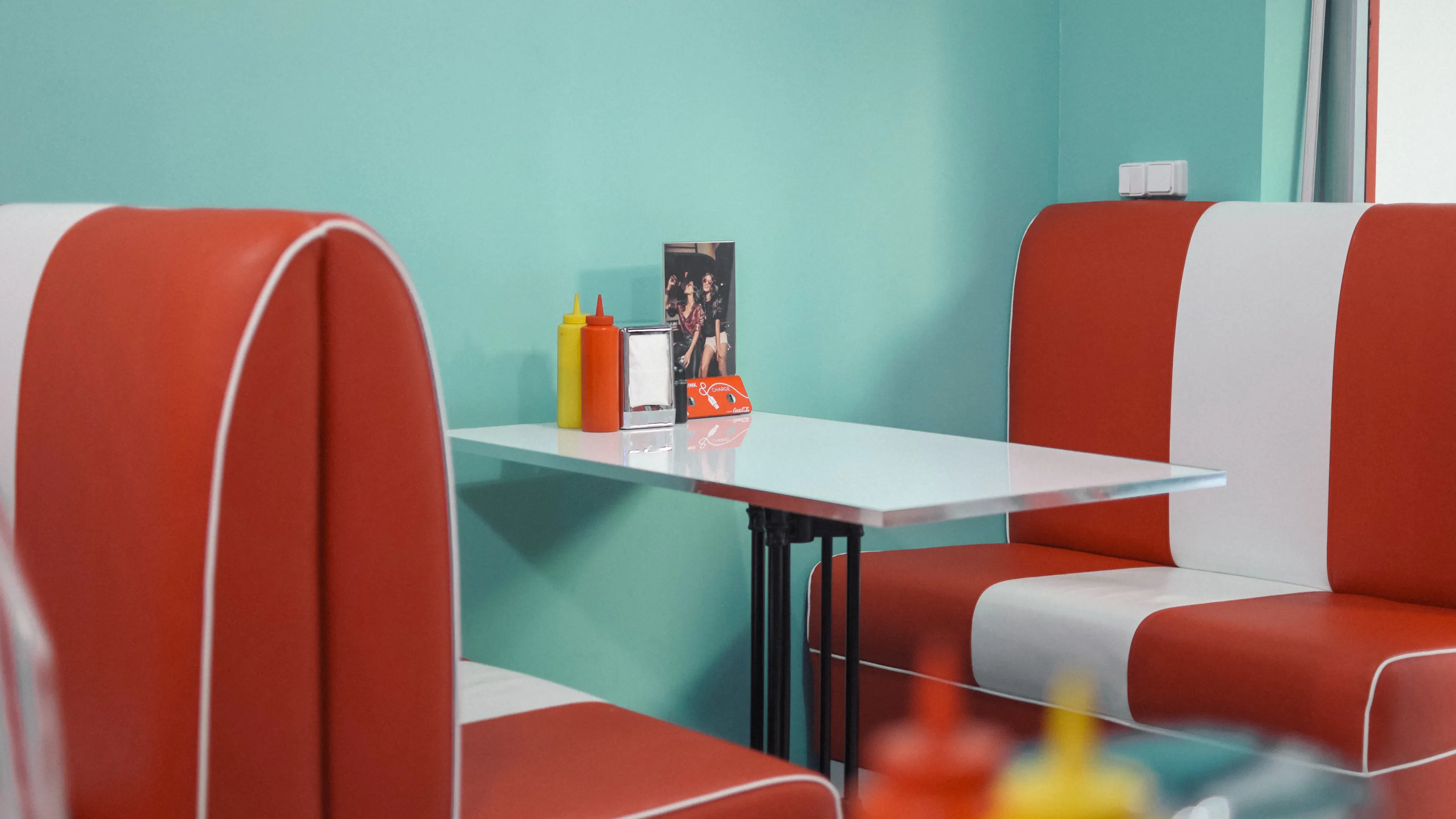 An empty booth with red and white leather seats at a retro diner with a turquoise wall and a bottle of ketchup and mustard on the table