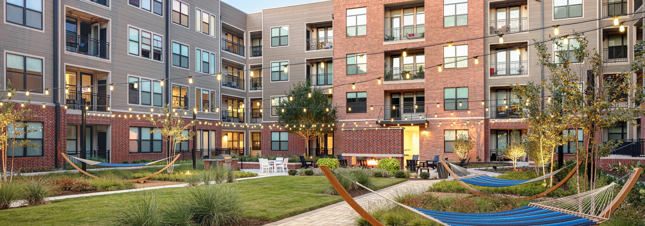 Dusk view of AMLI Addison grassy courtyard with striped hammocks and trees and other seating with suspended outdoor lights