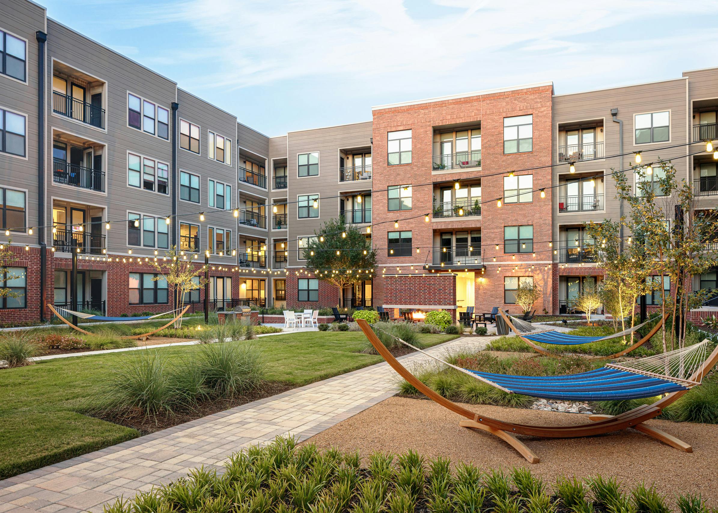 Dusk view of AMLI Addison grassy courtyard with striped hammocks and trees and other seating with suspended outdoor lights