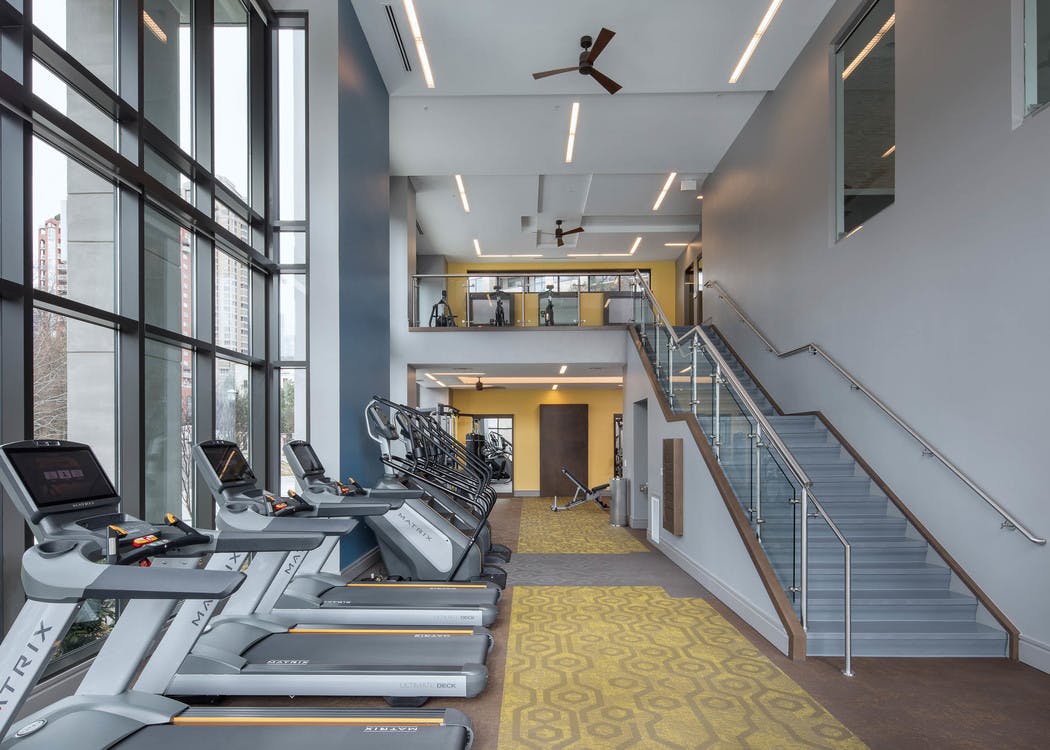 Fitness center at AMLI 3464 with yellow carpet and treadmills near tall windows with a modern staircase in the background