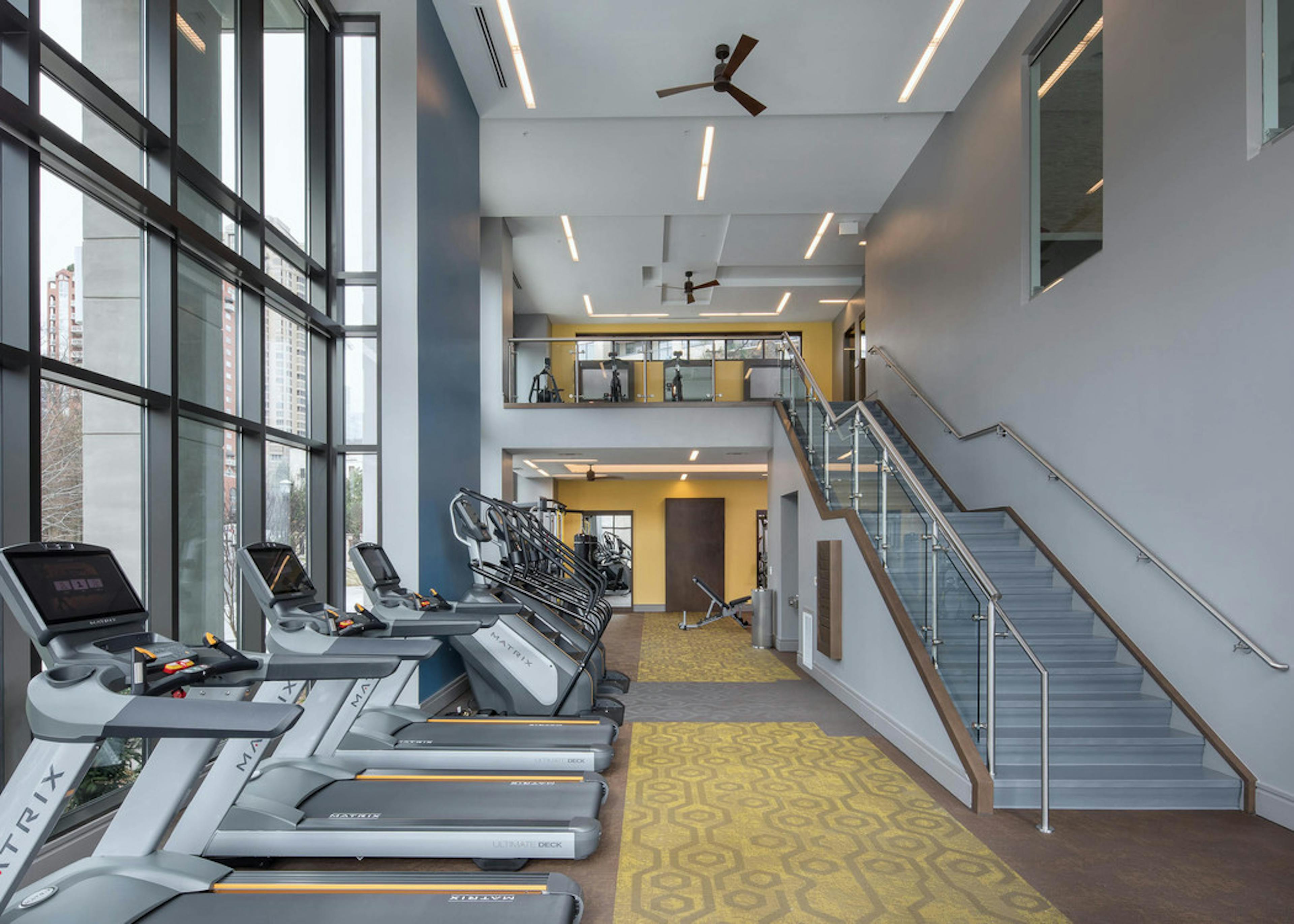 Fitness center at AMLI 3464 with yellow carpet and treadmills near tall windows with a modern staircase in the background