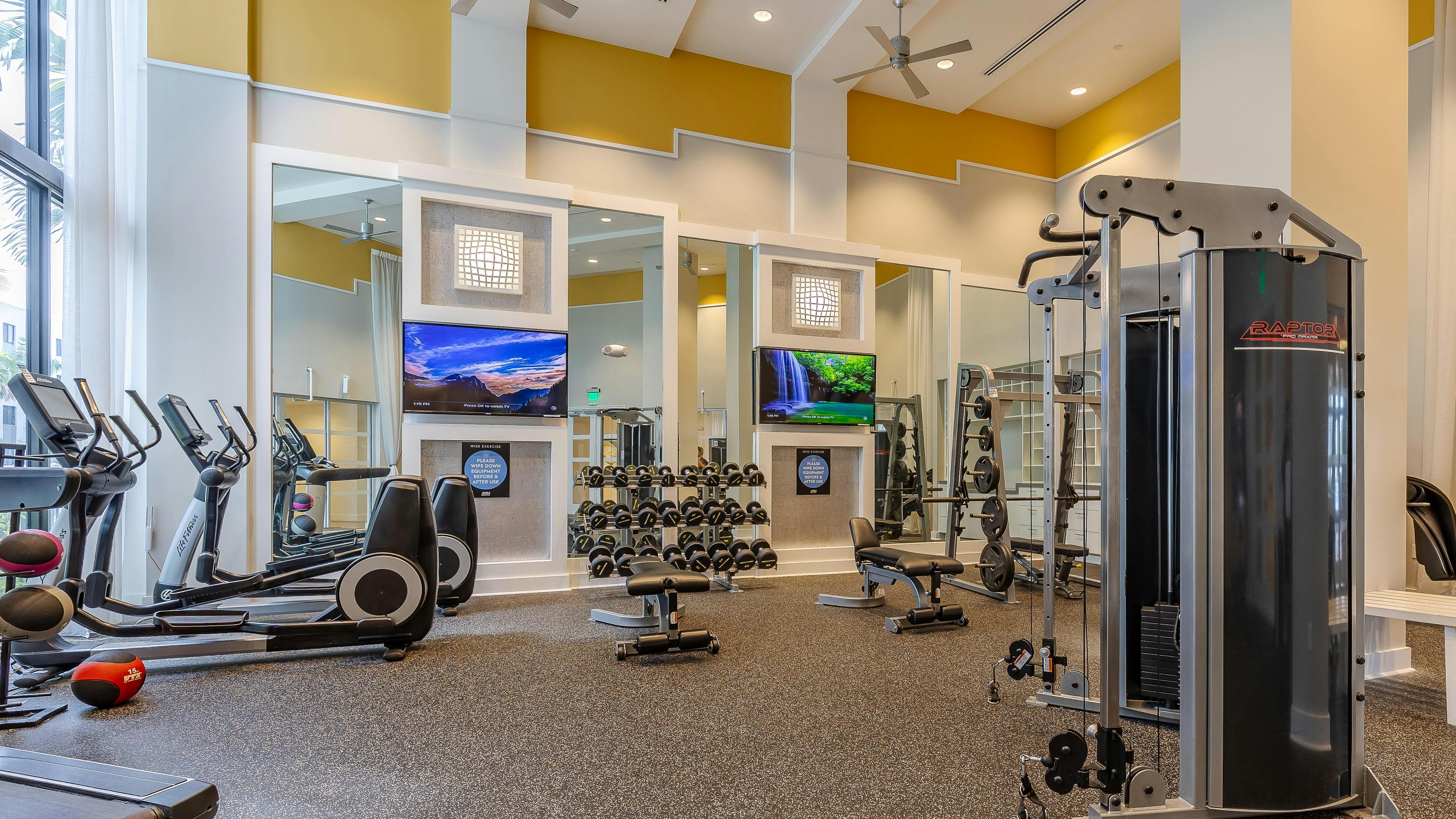 The fitness center at AMLI 8800 with free weights and cardio machines with TVs on the wall and tall ceilings with fans