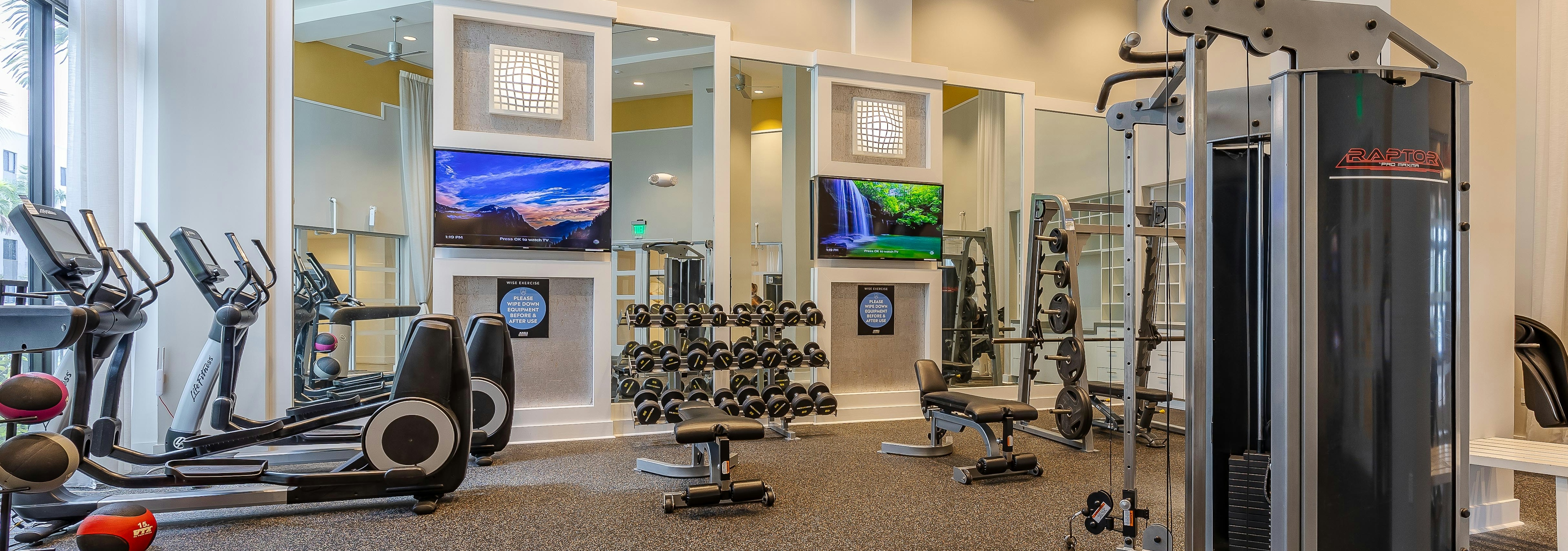 The fitness center at AMLI 8800 with free weights and cardio machines with TVs on the wall and tall ceilings with fans