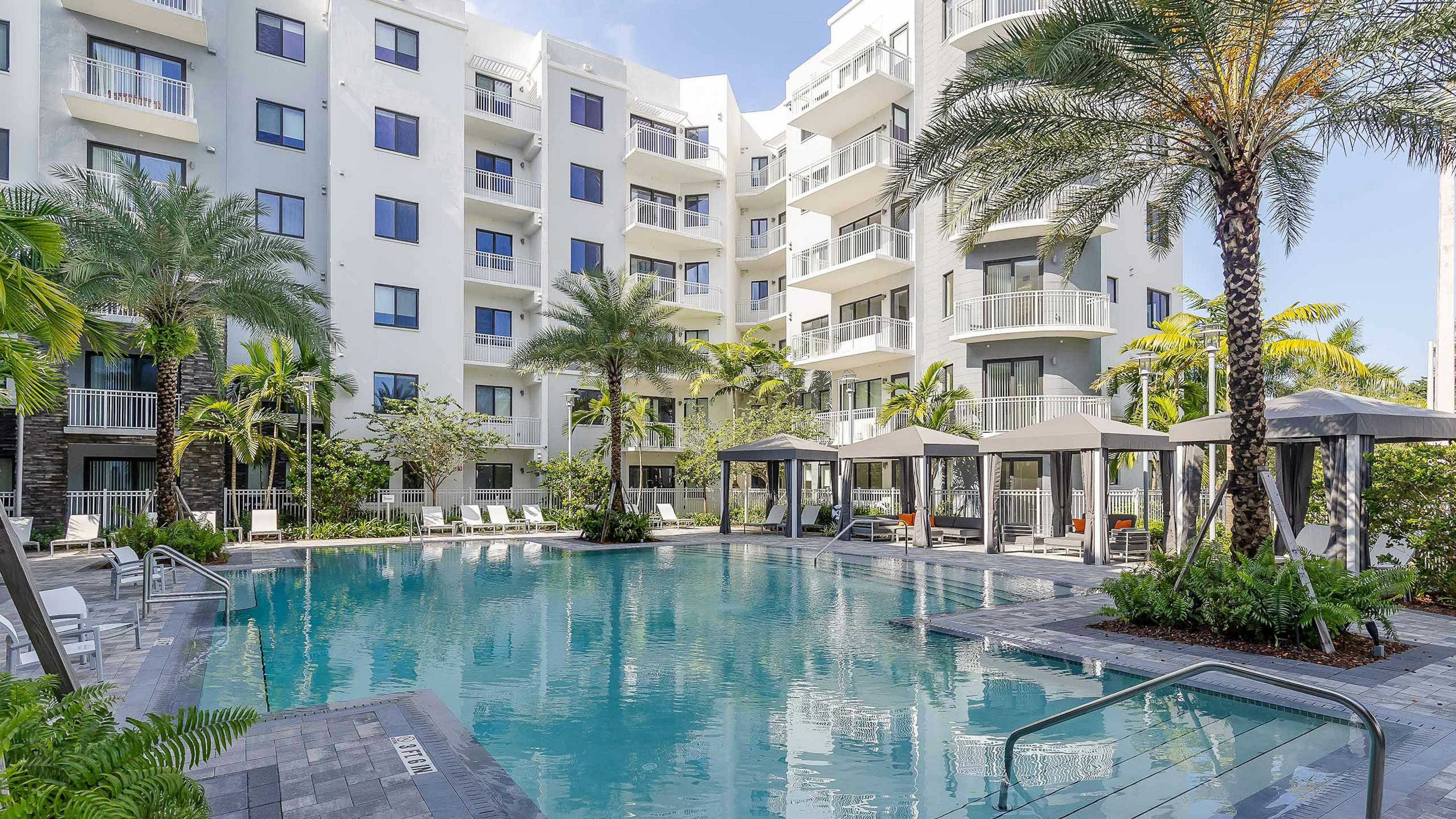 The pool at AMLI Park West apartments with cabanas and white lounge chairs with gray herringbone pavers and lush landscape 