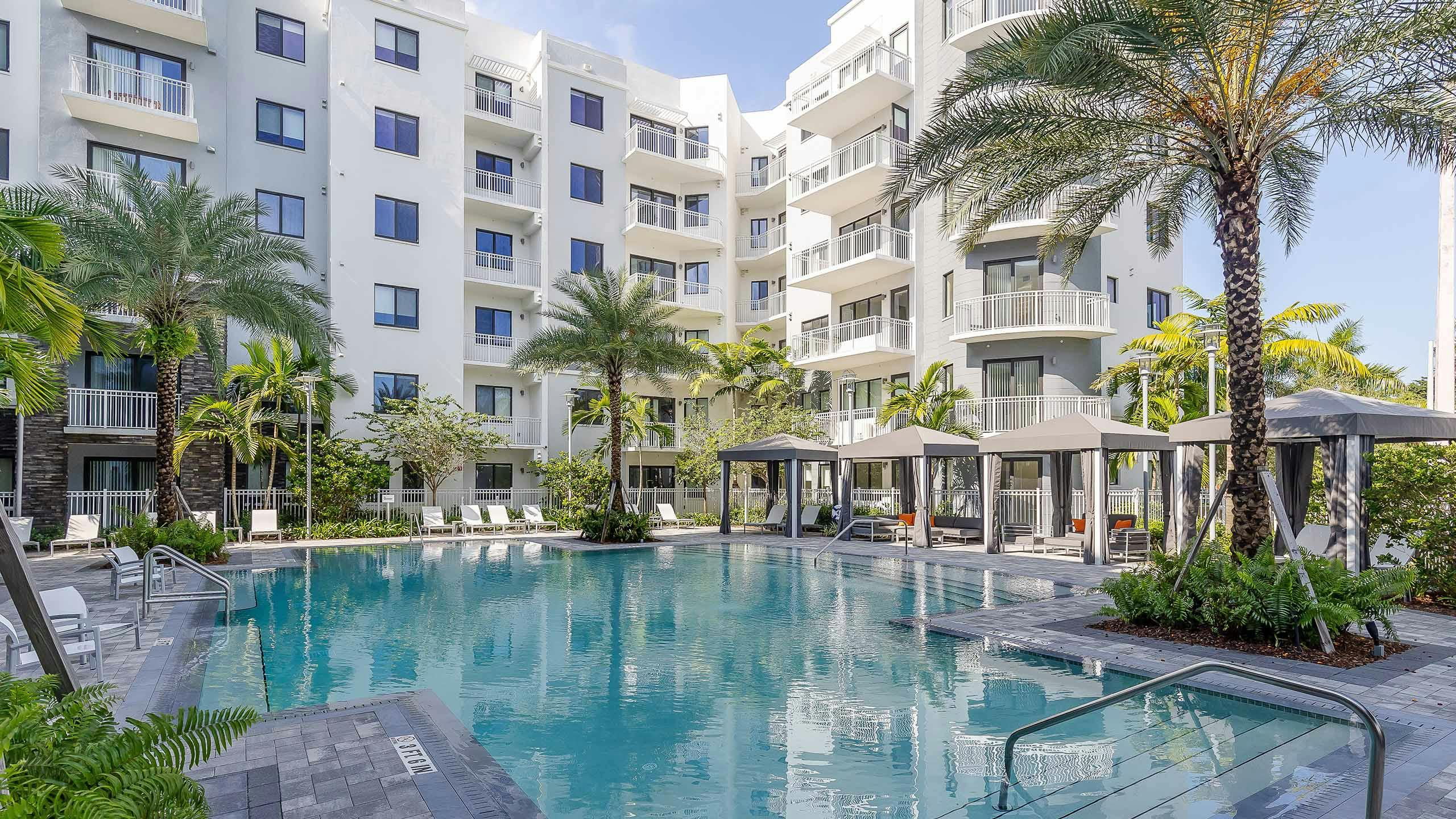The pool at AMLI Park West apartments with cabanas and white lounge chairs with gray herringbone pavers and lush landscape