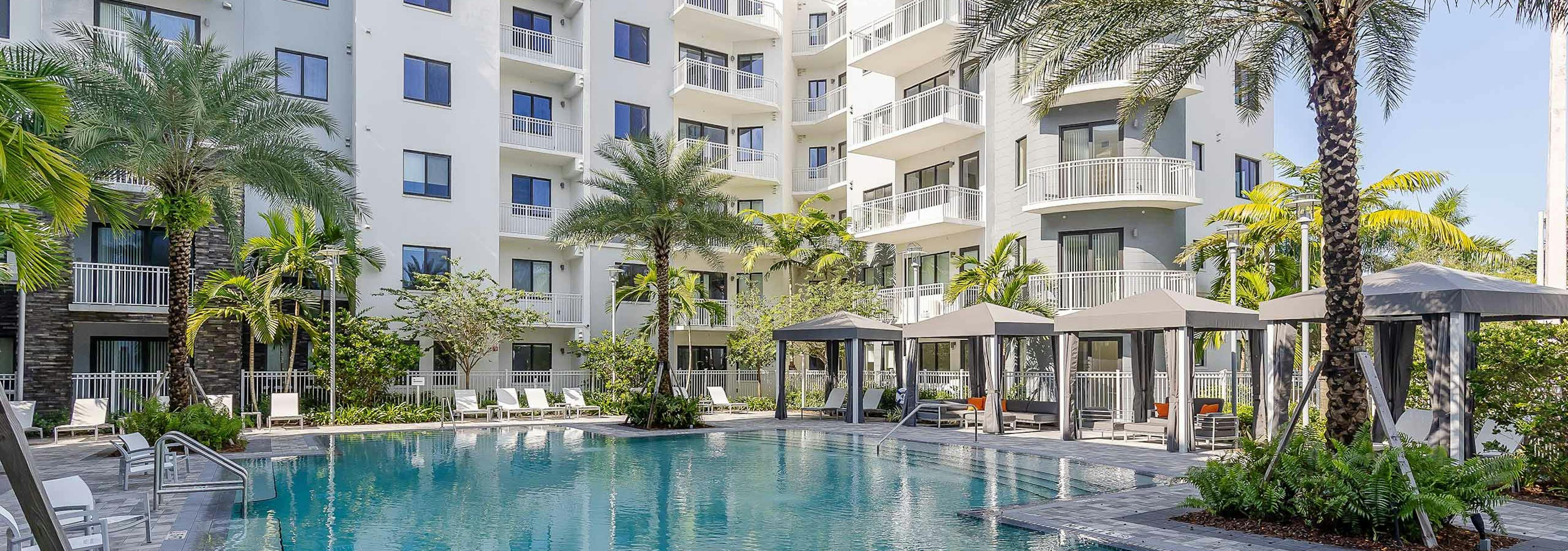 The pool at AMLI Park West apartments with cabanas and white lounge chairs with gray herringbone pavers and lush landscape 