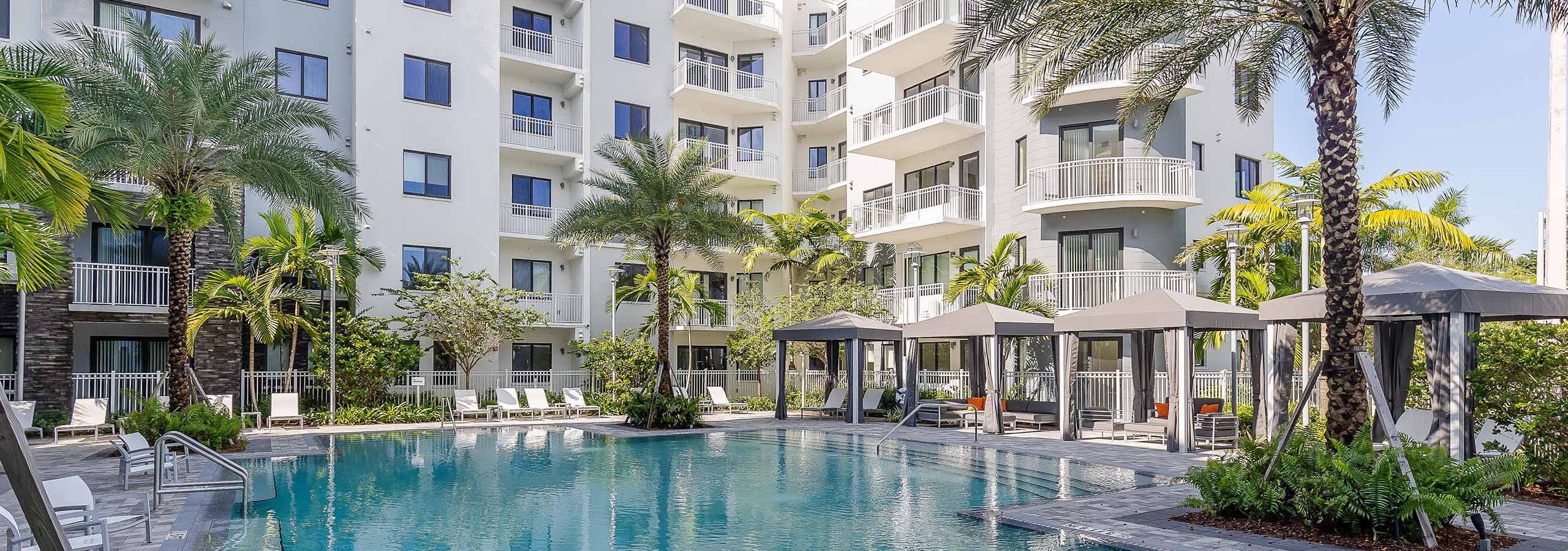 The pool at AMLI Park West apartments with cabanas and white lounge chairs with gray herringbone pavers and lush landscape