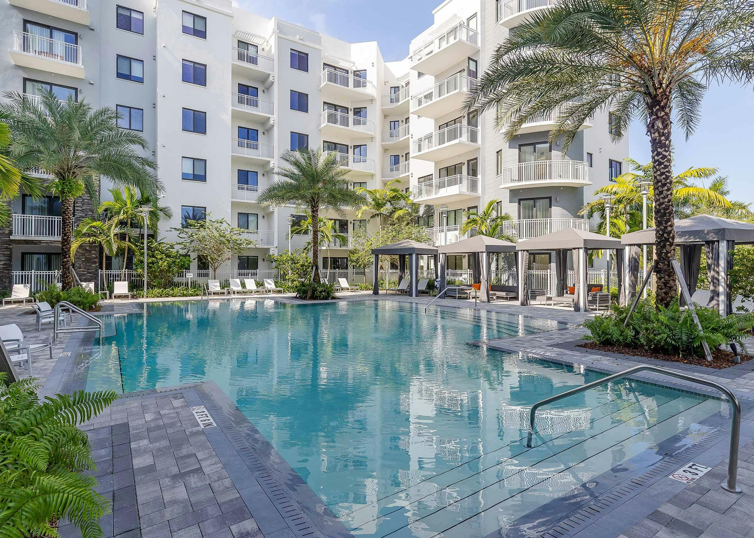 The pool at AMLI Park West apartments with cabanas and white lounge chairs with gray herringbone pavers and lush landscape