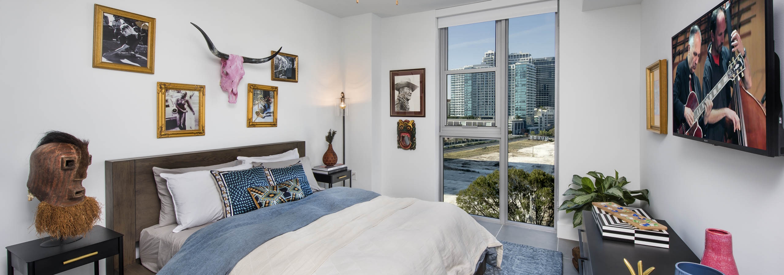 Bedroom with a city view and pink skull with horns over bed with white and blue blankets and blue carpet at AMLI Midtown 29