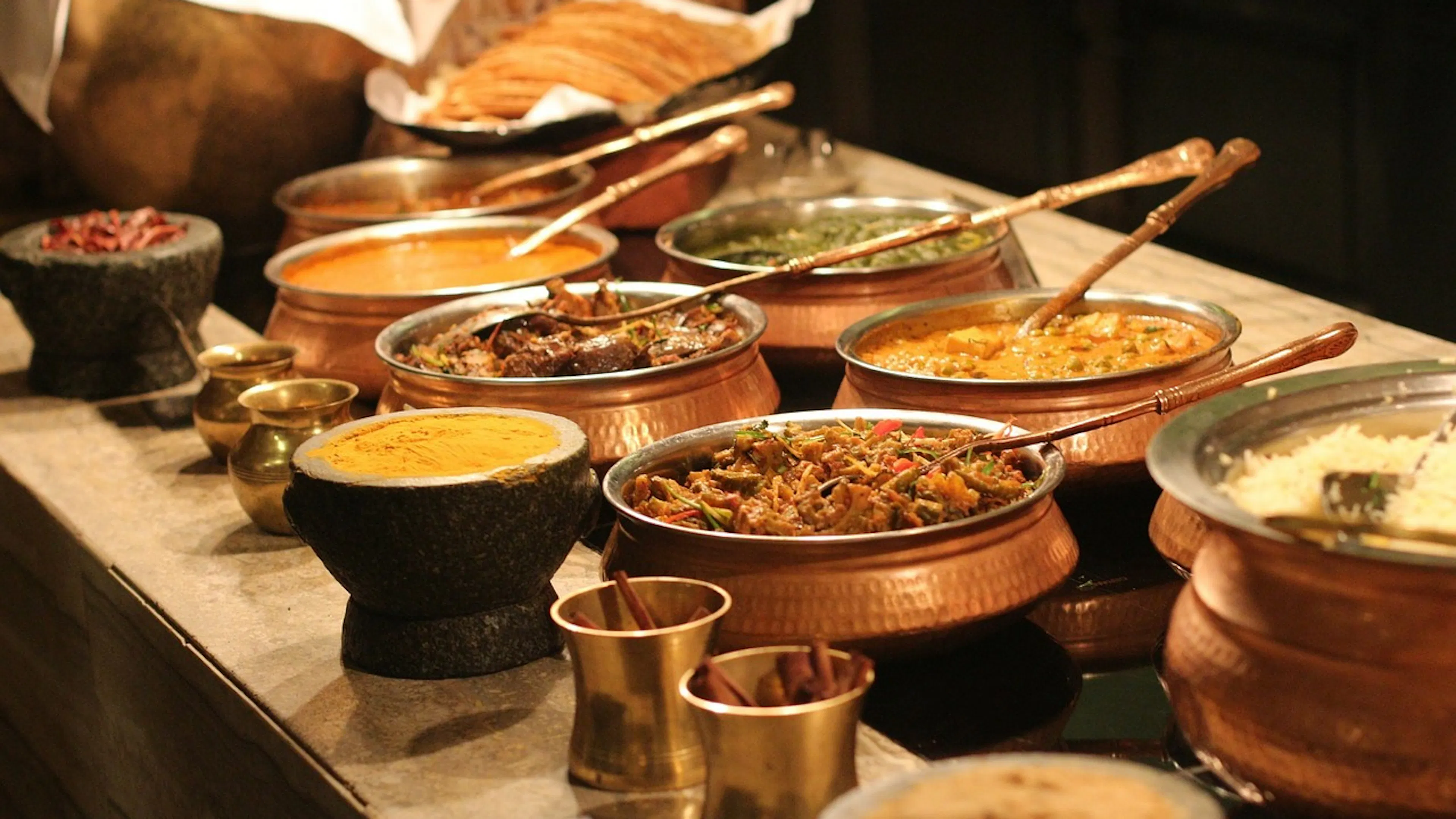 A dimly lit buffet table with 8 pots of curry rice, vegetables and other dishes and foods