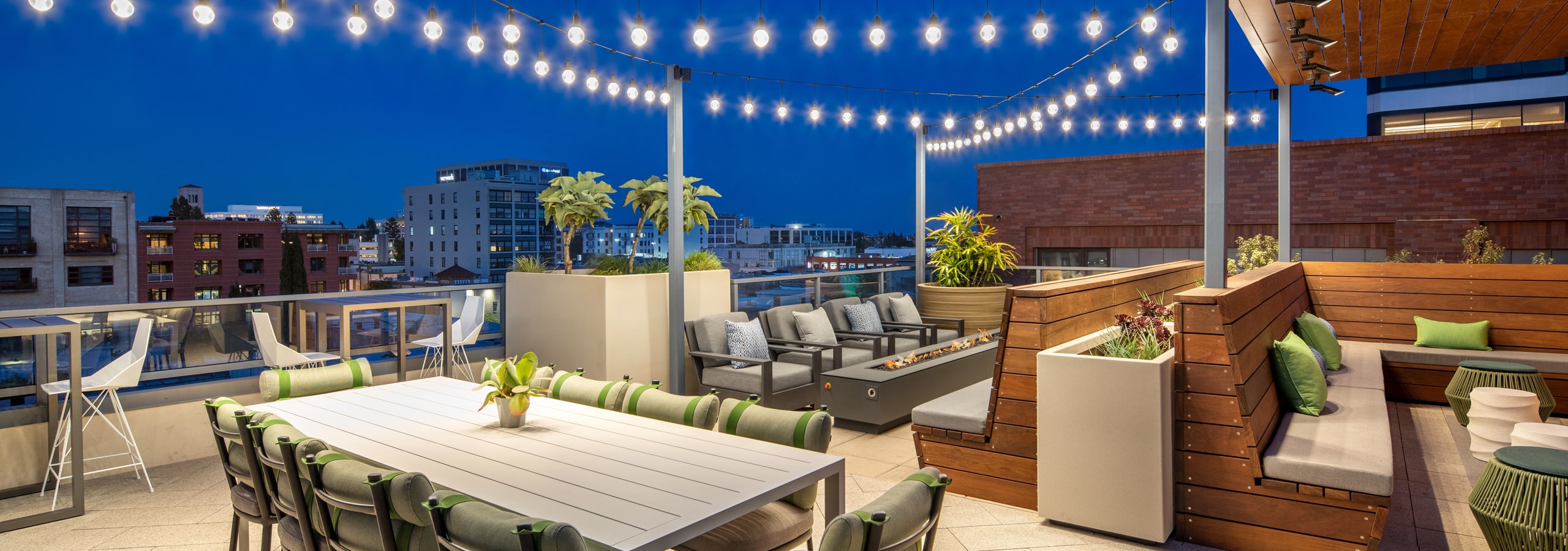 Nighttime view of rooftop sky deck with white table and cushioned chairs and firepit and hanging lights at AMLI Old Pasadena