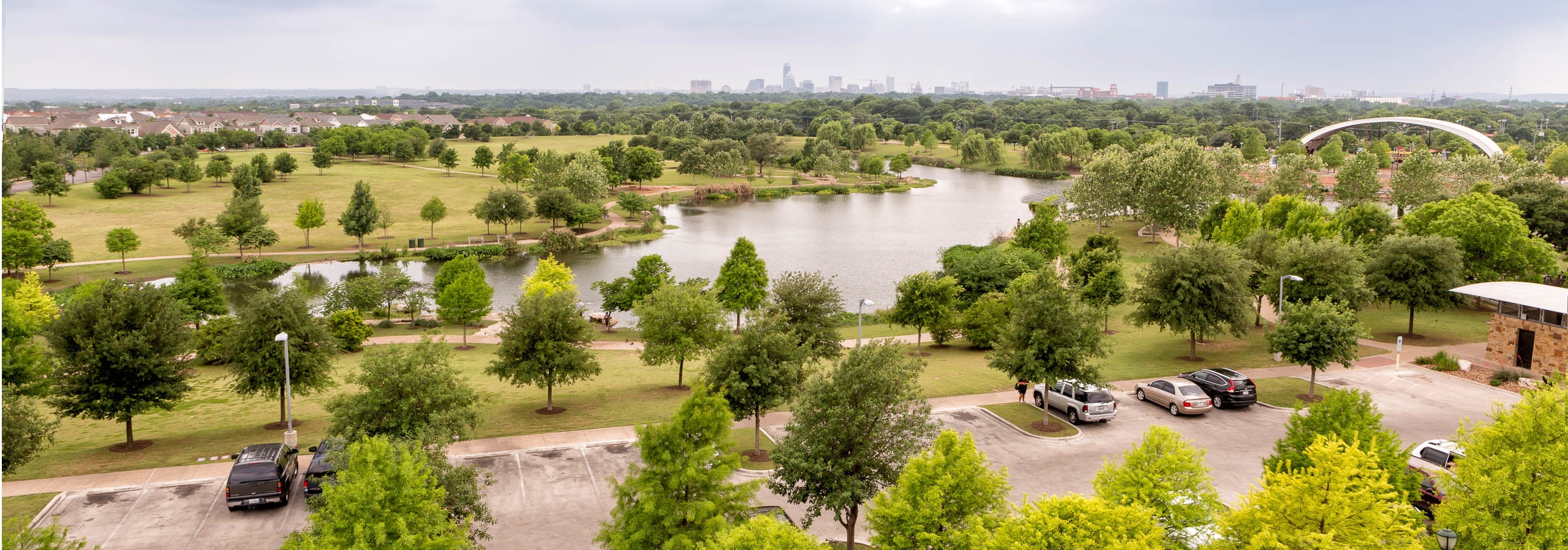 Overview of scattered vibrant green trees under a blue sky surrounding a grassy lake area with cars parked in a nearby lot